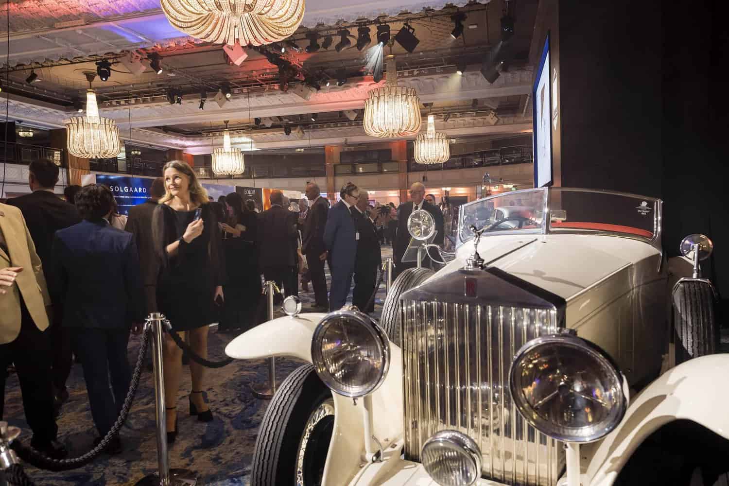 Strive for perfection while guests mingle near a classic white vintage car under chandeliers in an elegantly adorned venue.