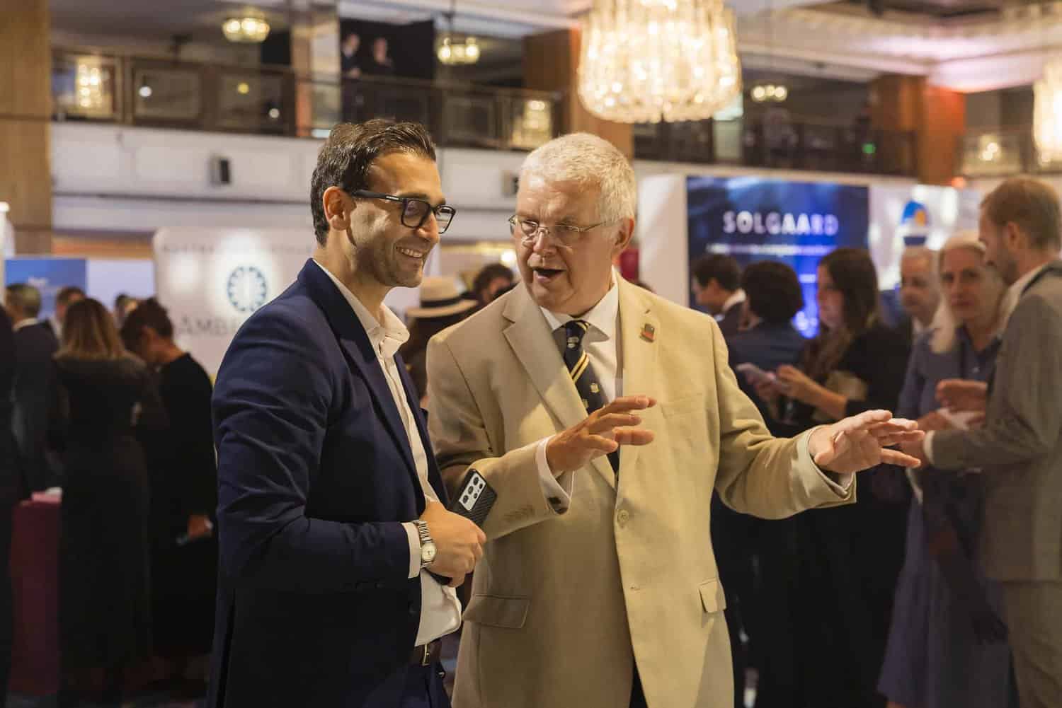 Two men engaged in conversation at an indoor event, surrounded by people and exhibition stands, embodying the spirit to strive for perfection in every interaction.
