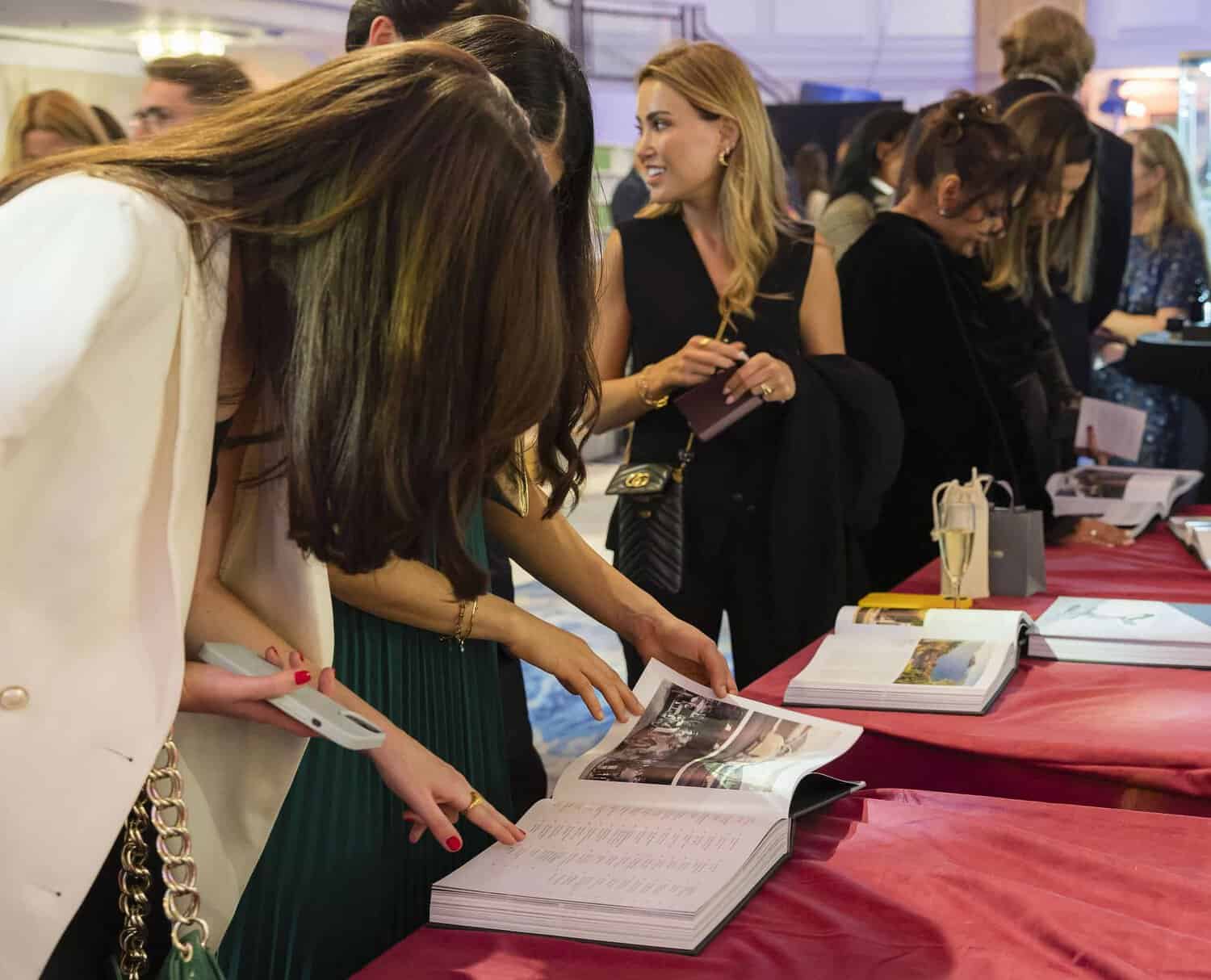People striving for perfection gather around an open book on a red table, engaging in spirited discussion while surrounded by various intriguing items.