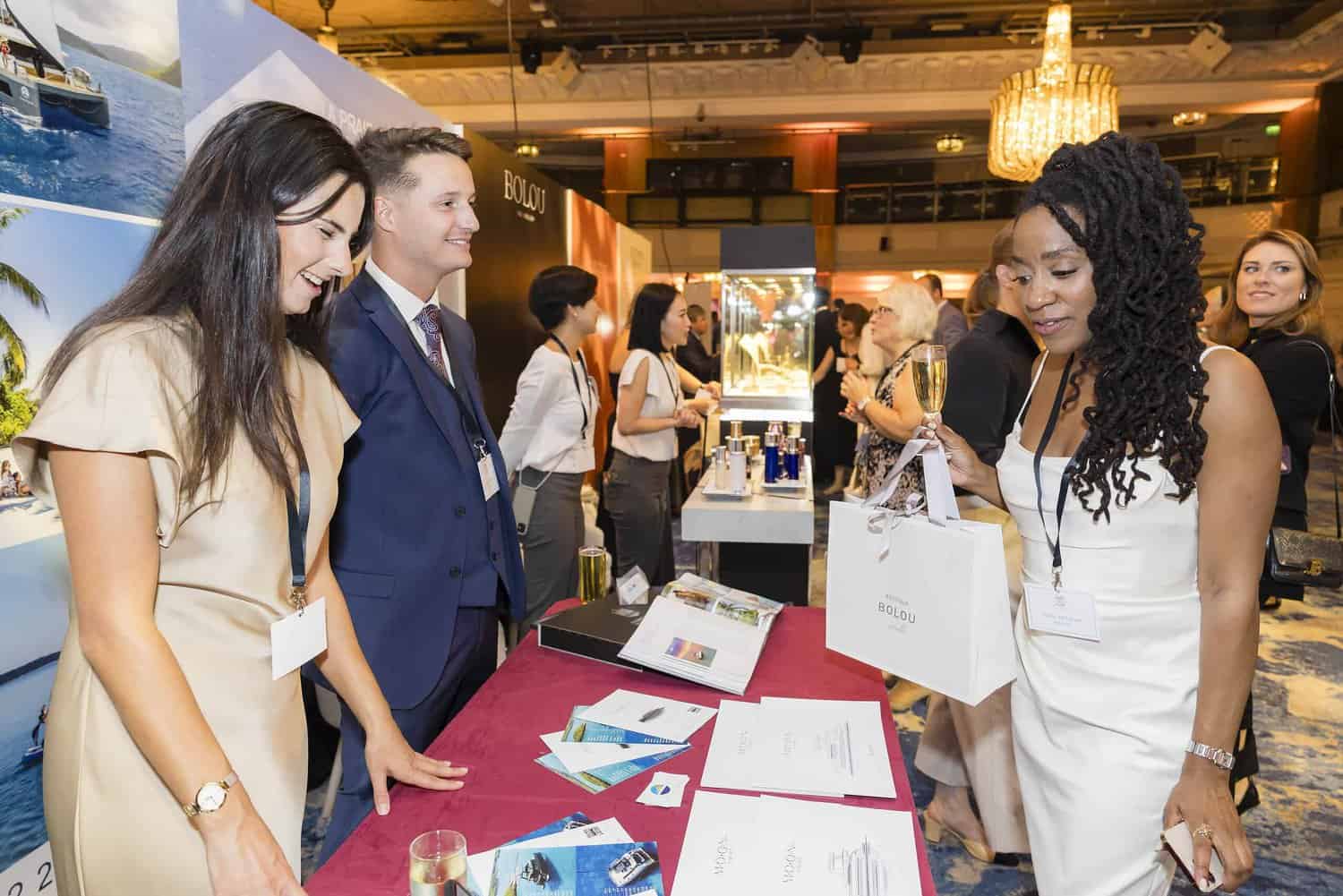 At a bustling trade show booth, two presenters stand behind a table lined with brochures, expertly discussing their offerings. A visitor, holding a shopping bag and glass, engages them in conversation, captivated by the seamless presentation—an embodiment of trade show perfection.