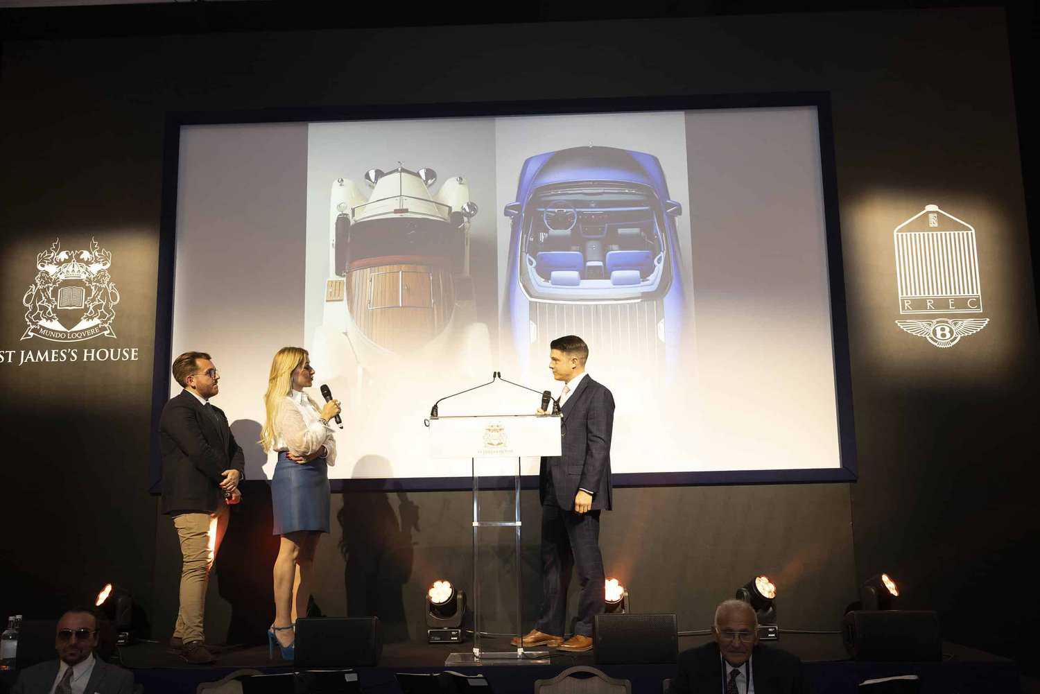Three people stand on stage with microphones, embodying a strive for perfection, in front of a large presentation screen displaying stunning images of luxury cars viewed from above.