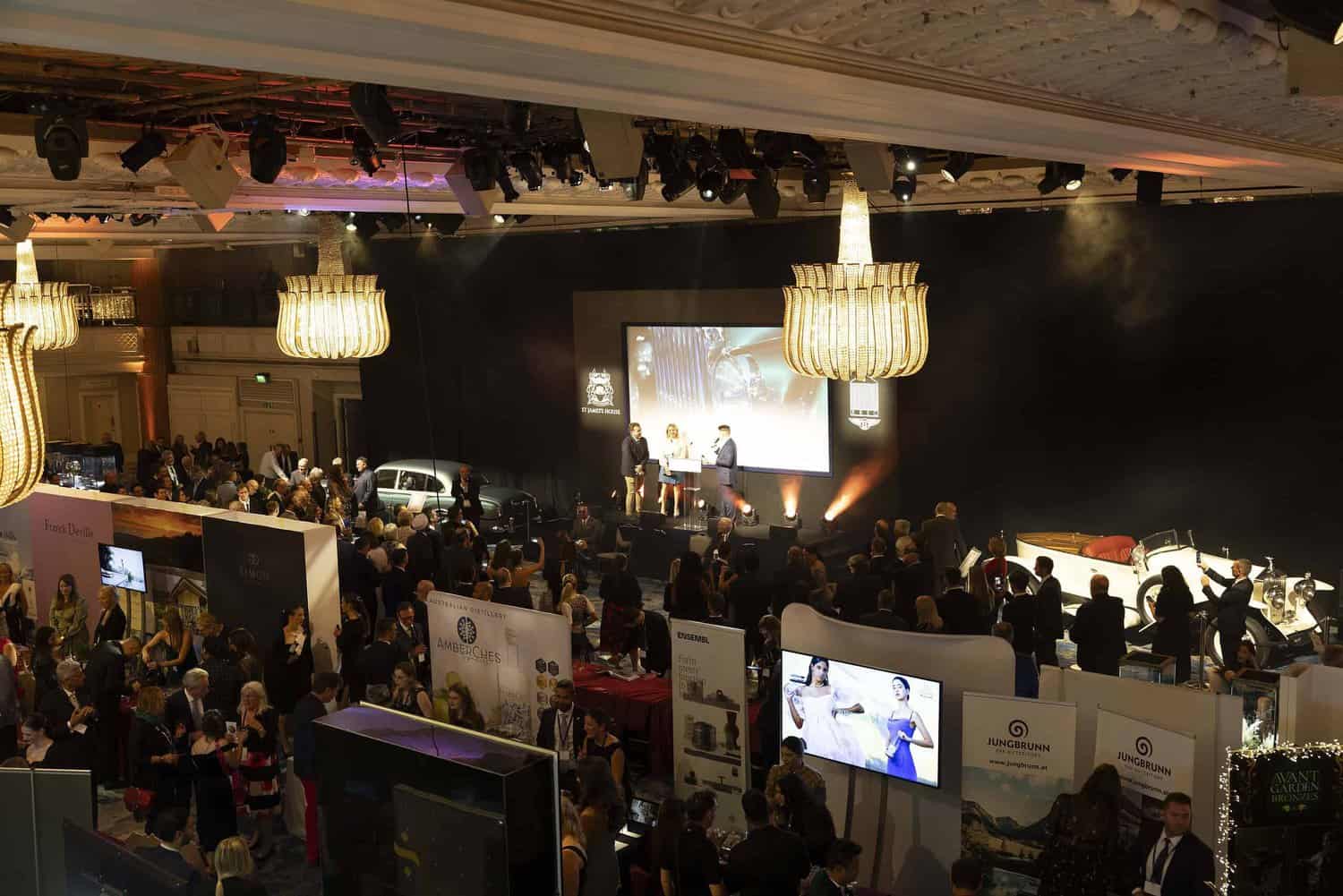 A bustling event with numerous booths, people interacting, and a stage where speakers present under ornate chandeliers in a grand hall, creating an atmosphere of absolute perfection.