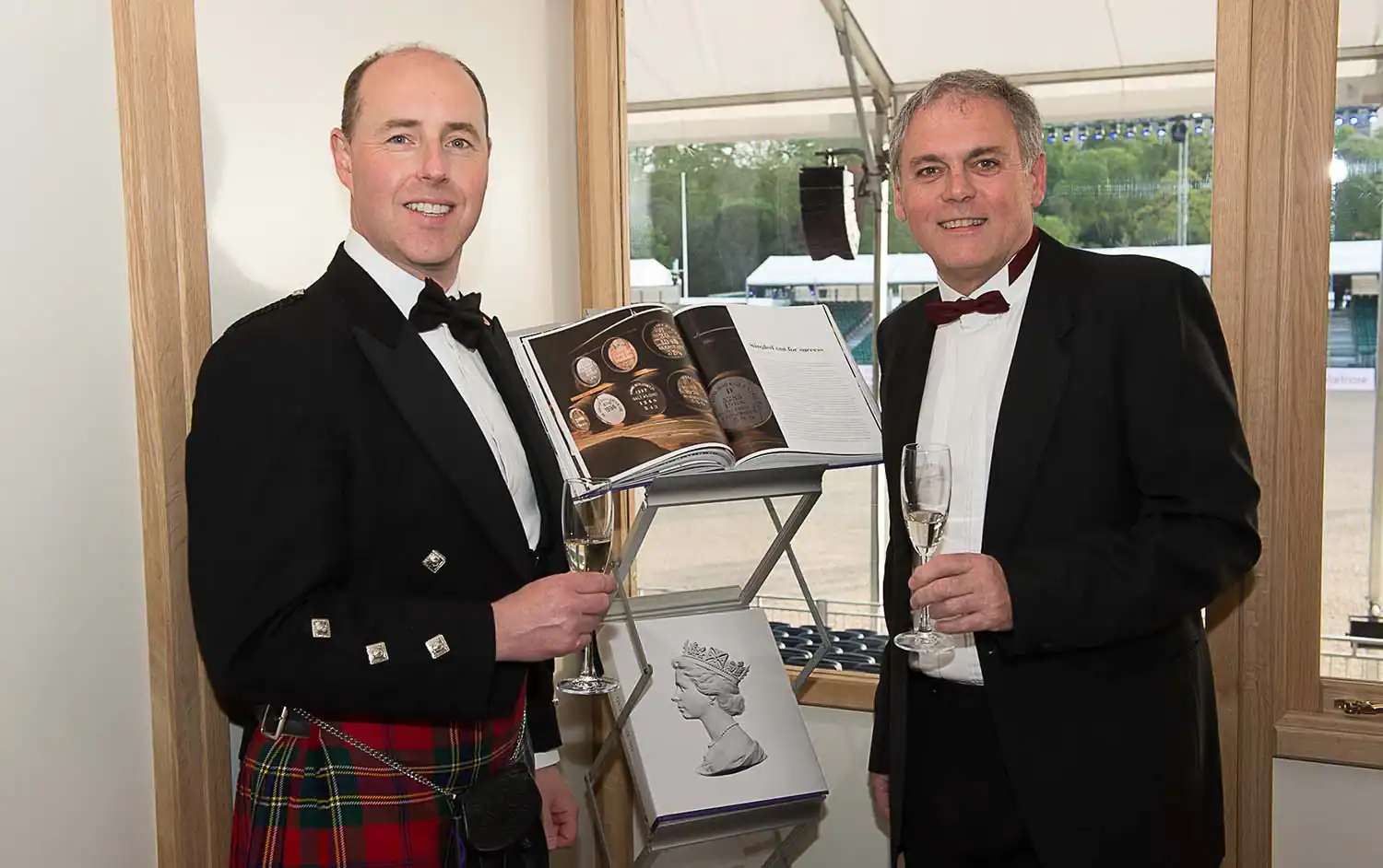 Two men in formal attire hold champagne glasses beside a display of coins and a book. One man wears a kilt, and there is a sketch of The Queen at 90 on the podium, celebrating her remarkable legacy.
