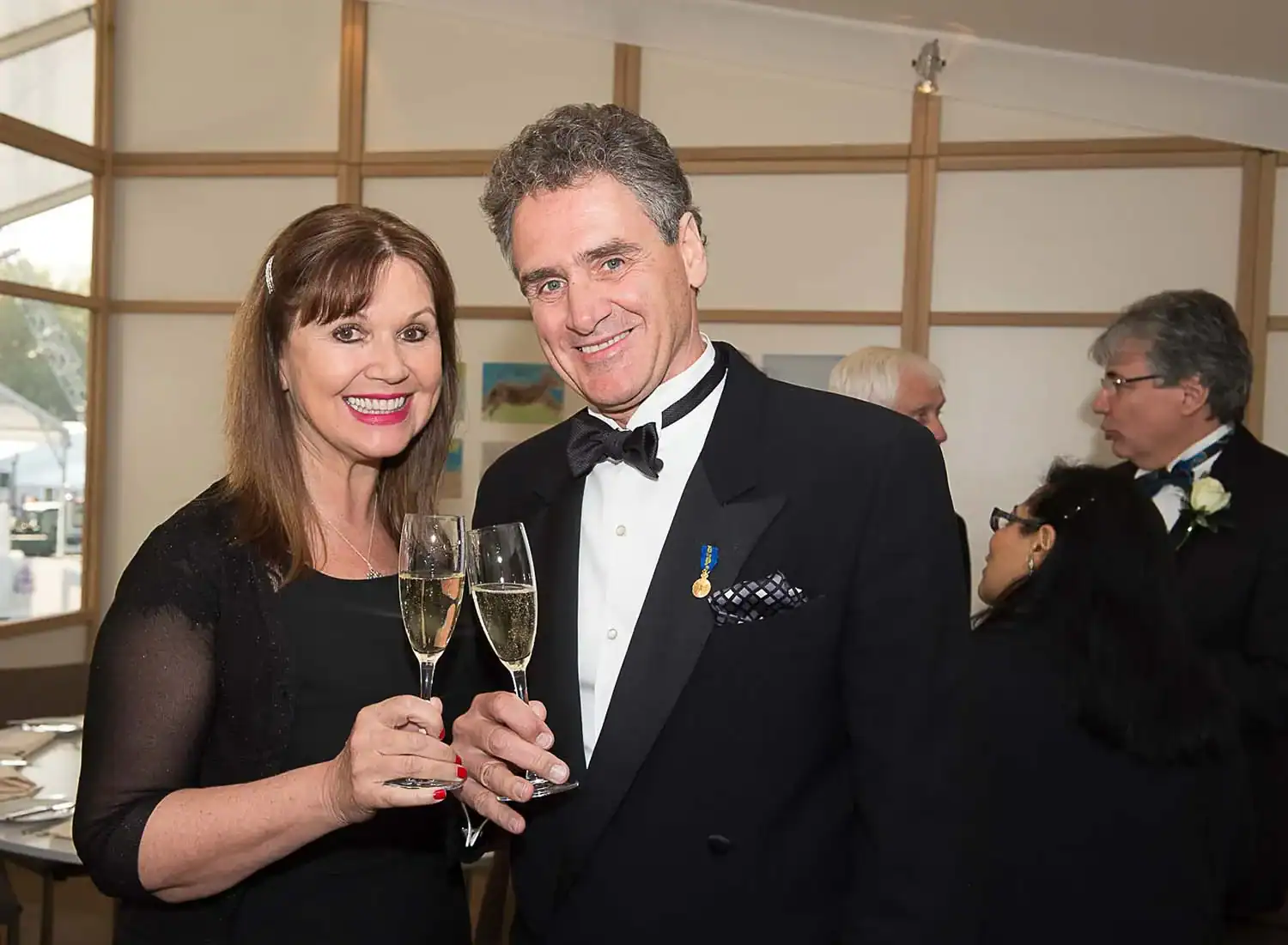 A man and woman in formal attire smile and hold champagne glasses at an indoor event, reminiscent of the elegance celebrated during The Queen at 90 festivities. Other guests are visible in the background.