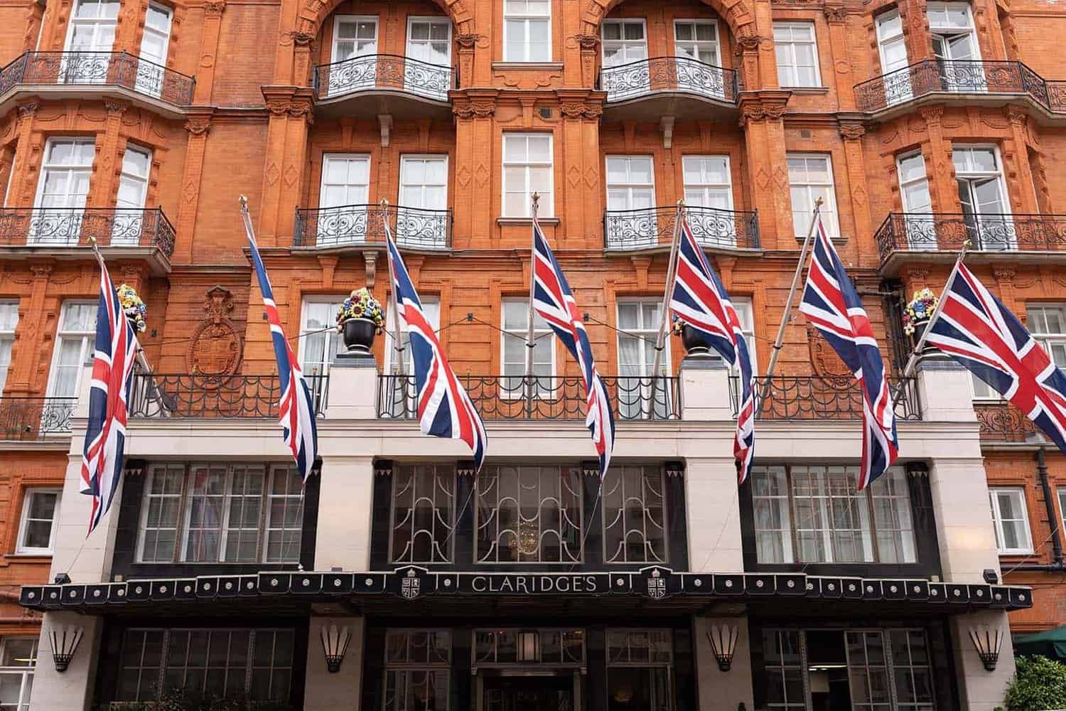 Red brick building facade with multiple Union Jack flags evokes a sense of royalty. Sign reads "Claridge's," where Her Majesty might feel at home. Balconies and ornate decorations grace the entrance.
