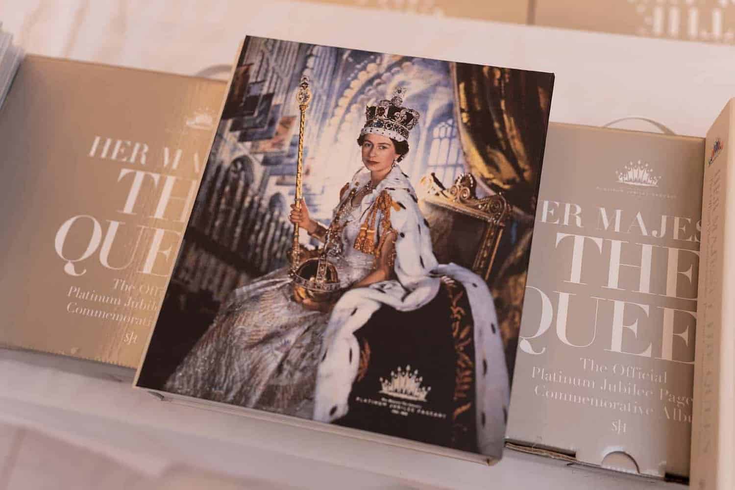 A commemorative book cover features a formal portrait of Her Majesty in regal attire and a crown, seated on a throne, holding a scepter.