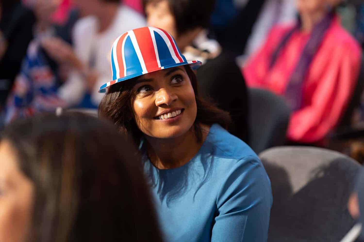 A woman wearing a blue shirt and a Union Jack hat, smiling with regal charm, is seated in the jubilant crowd celebrating Her Majesty.