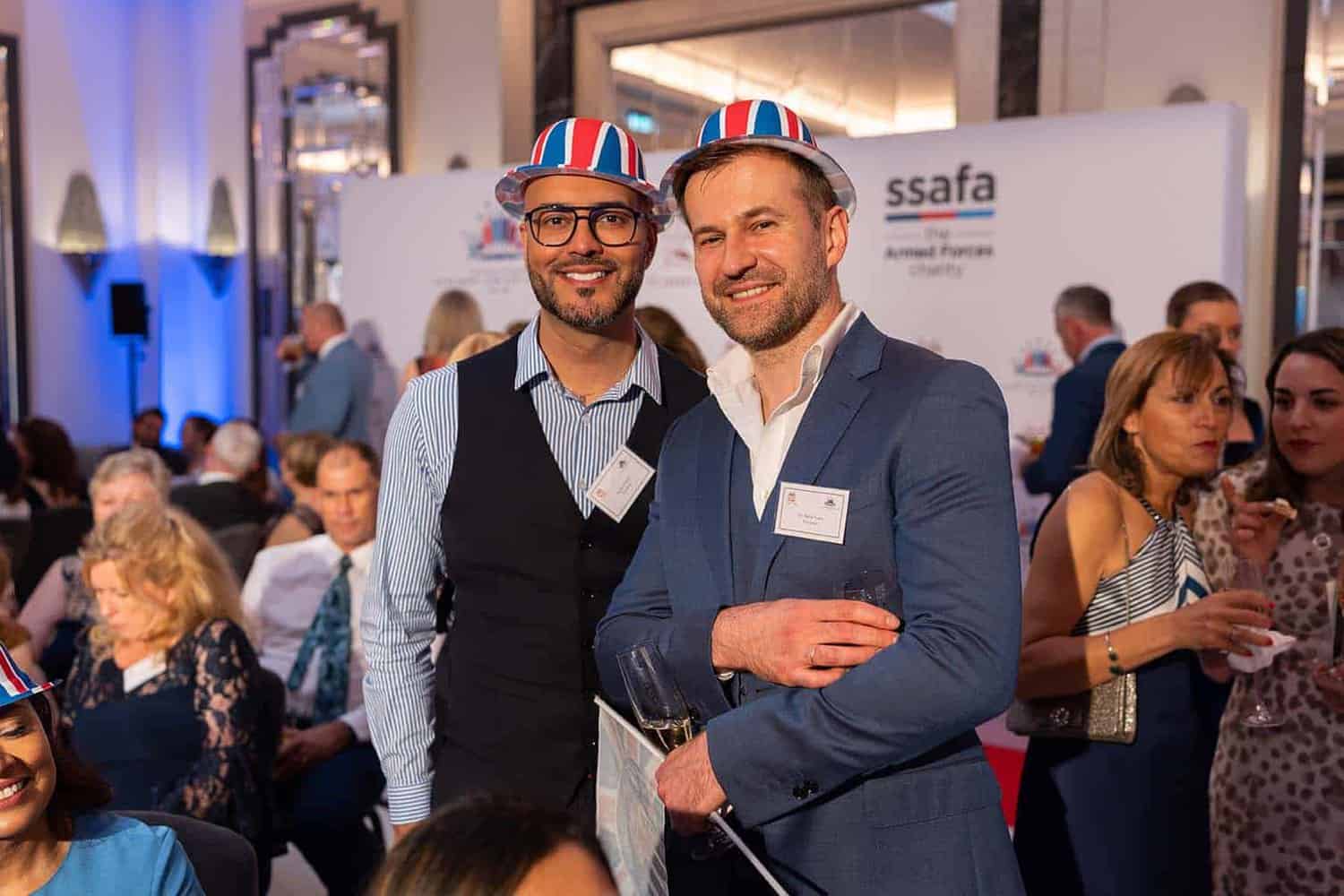Two men wearing ribbon hats stand together at an event with people in the background, a nod to Royalty seen through the SSAFA charity banner that honors service.