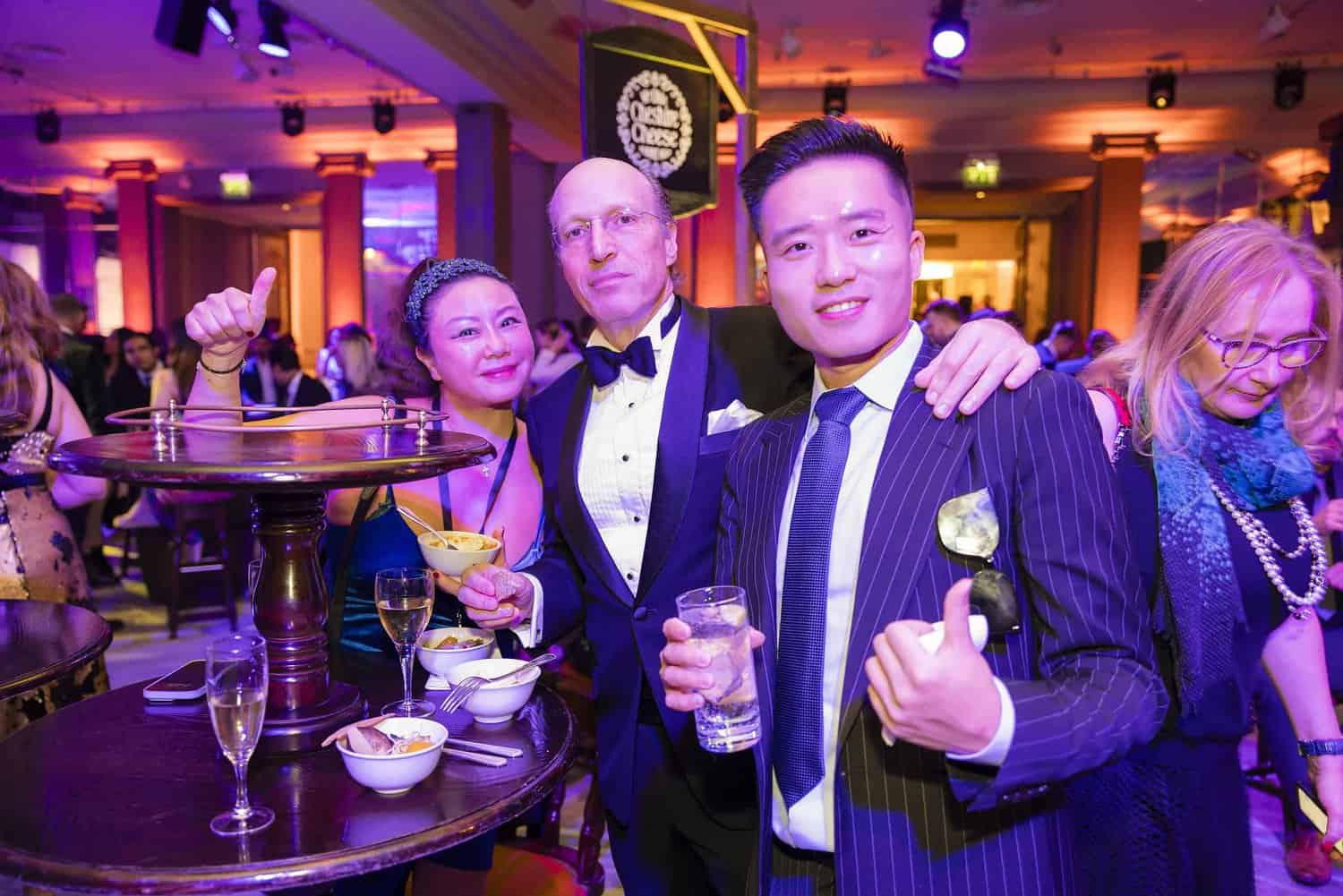 A group of people in formal attire are smiling and posing at a lively London party, with drinks and snacks on the table, reminiscent of a glamorous ball.