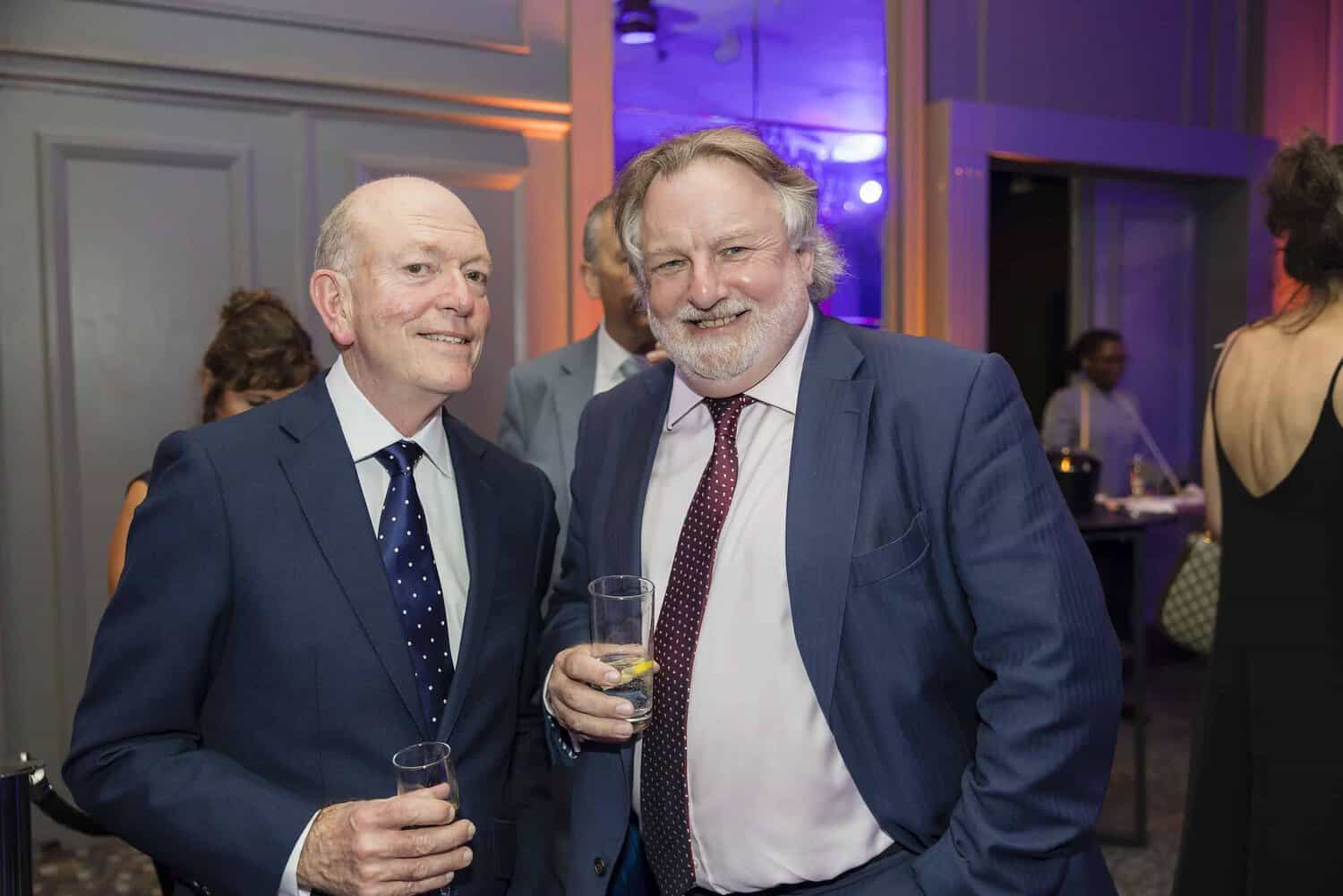 Two men in suits are smiling and holding drinks at the London Press Club Ball, enjoying the vibrant atmosphere of this splendid indoor event.
