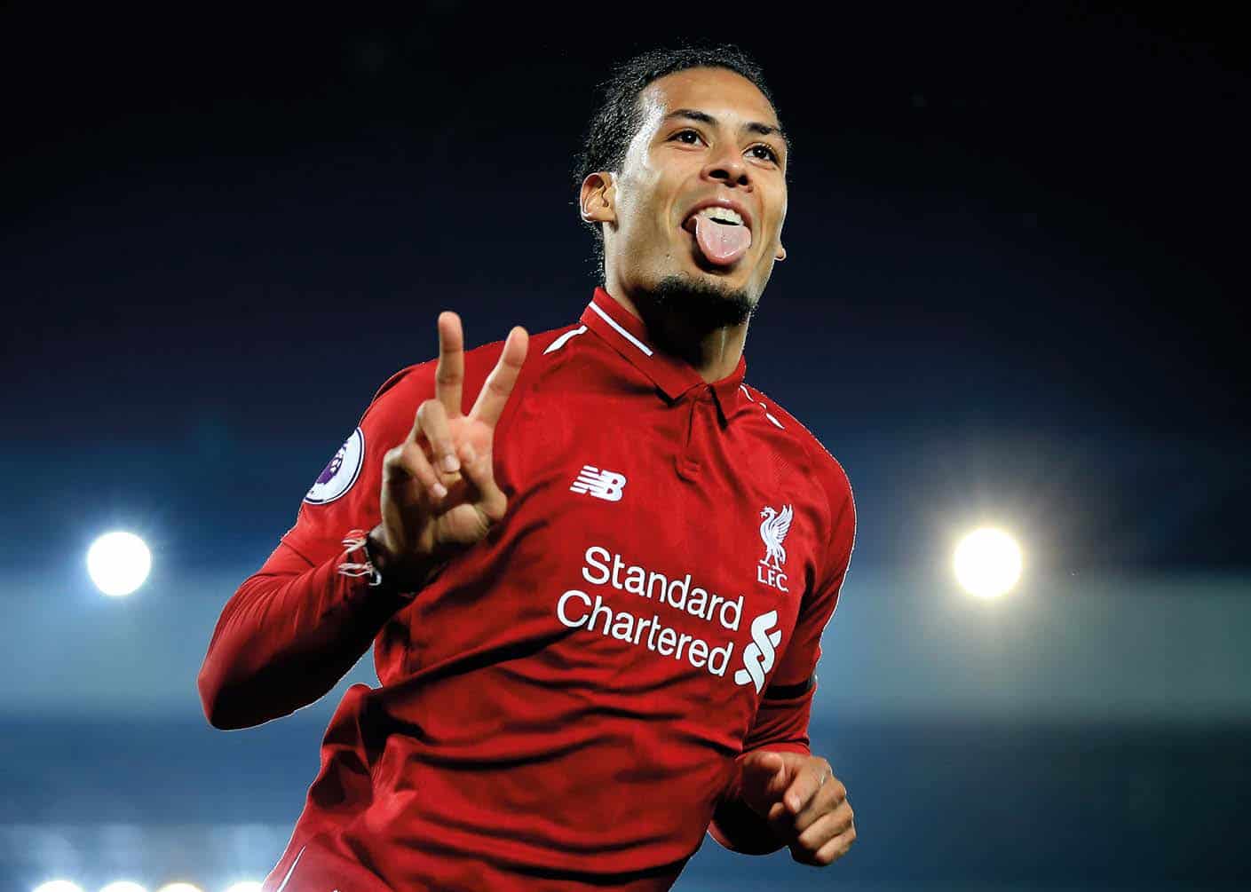 A Liverpool FC player in a red jersey with "Standard Chartered" gestures with a peace sign and sticks out his tongue on the field during the 2018/19 season.
