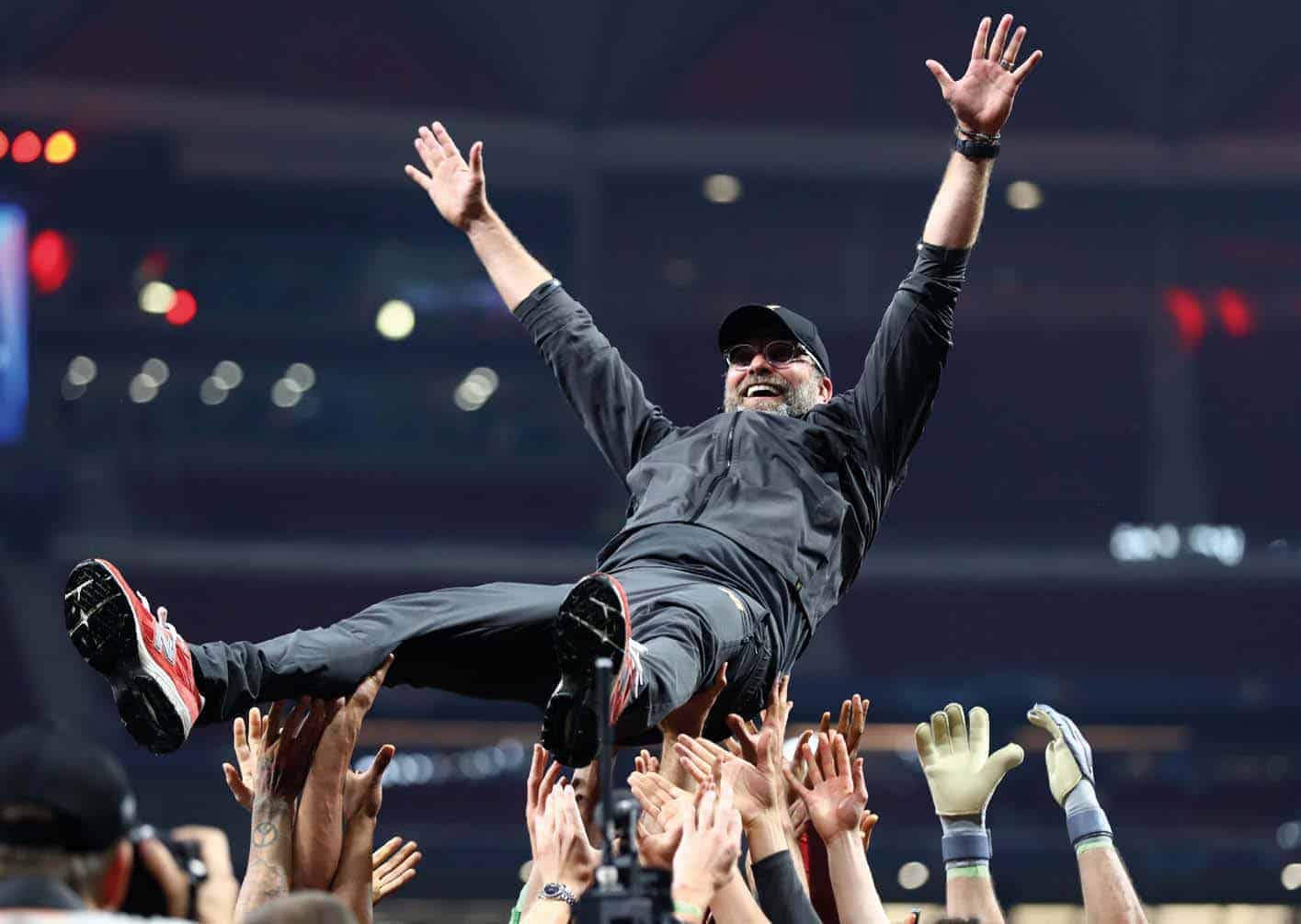 A person in sports attire and sunglasses is being lifted by a jubilant crowd under the bright lights of a stadium, celebrating a thrilling moment from Liverpool FC's unforgettable 2018/19 season.