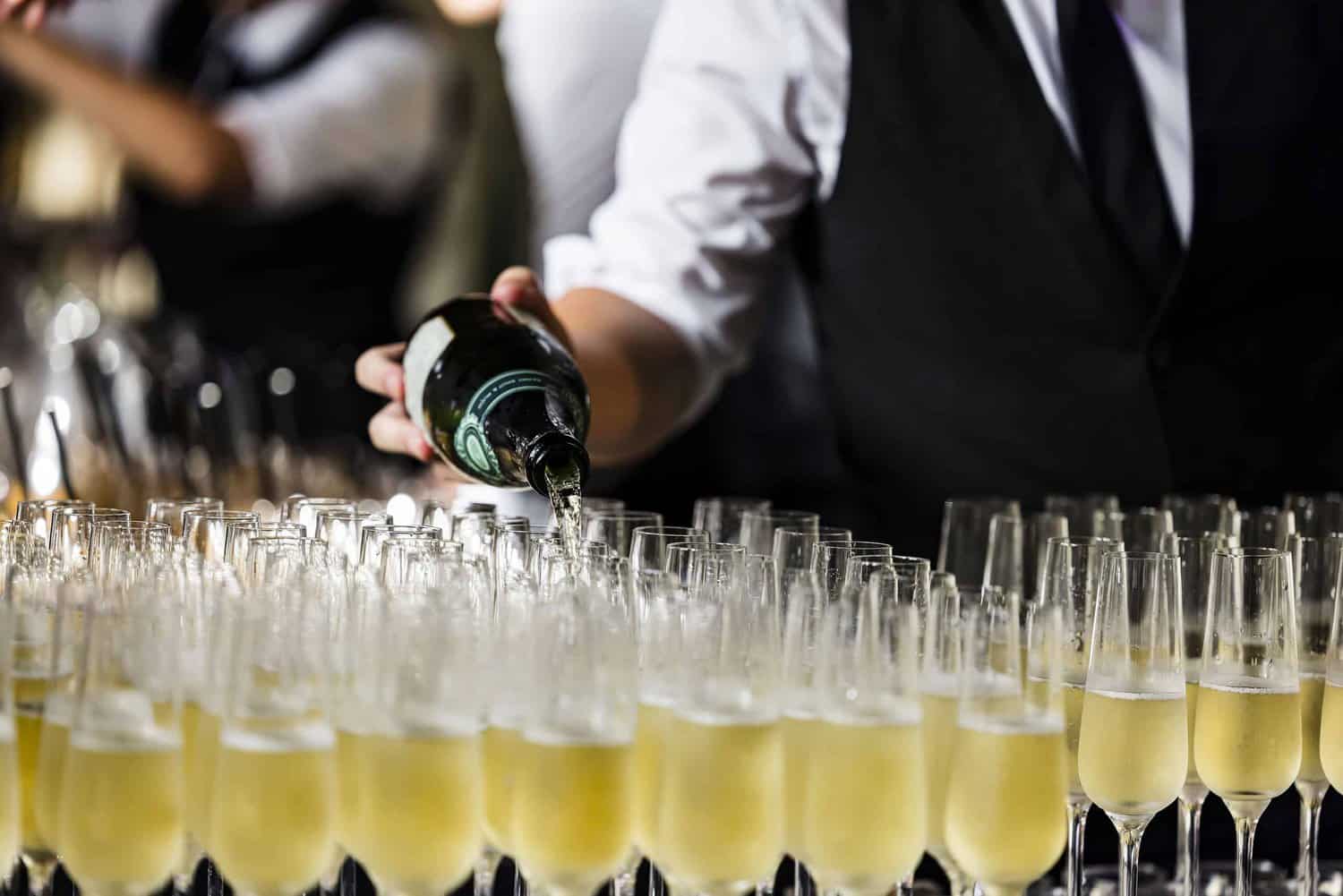 A person in formal attire pours champagne into a row of filled glasses, celebrating 75 years of the National Health Service with elegance and grace.