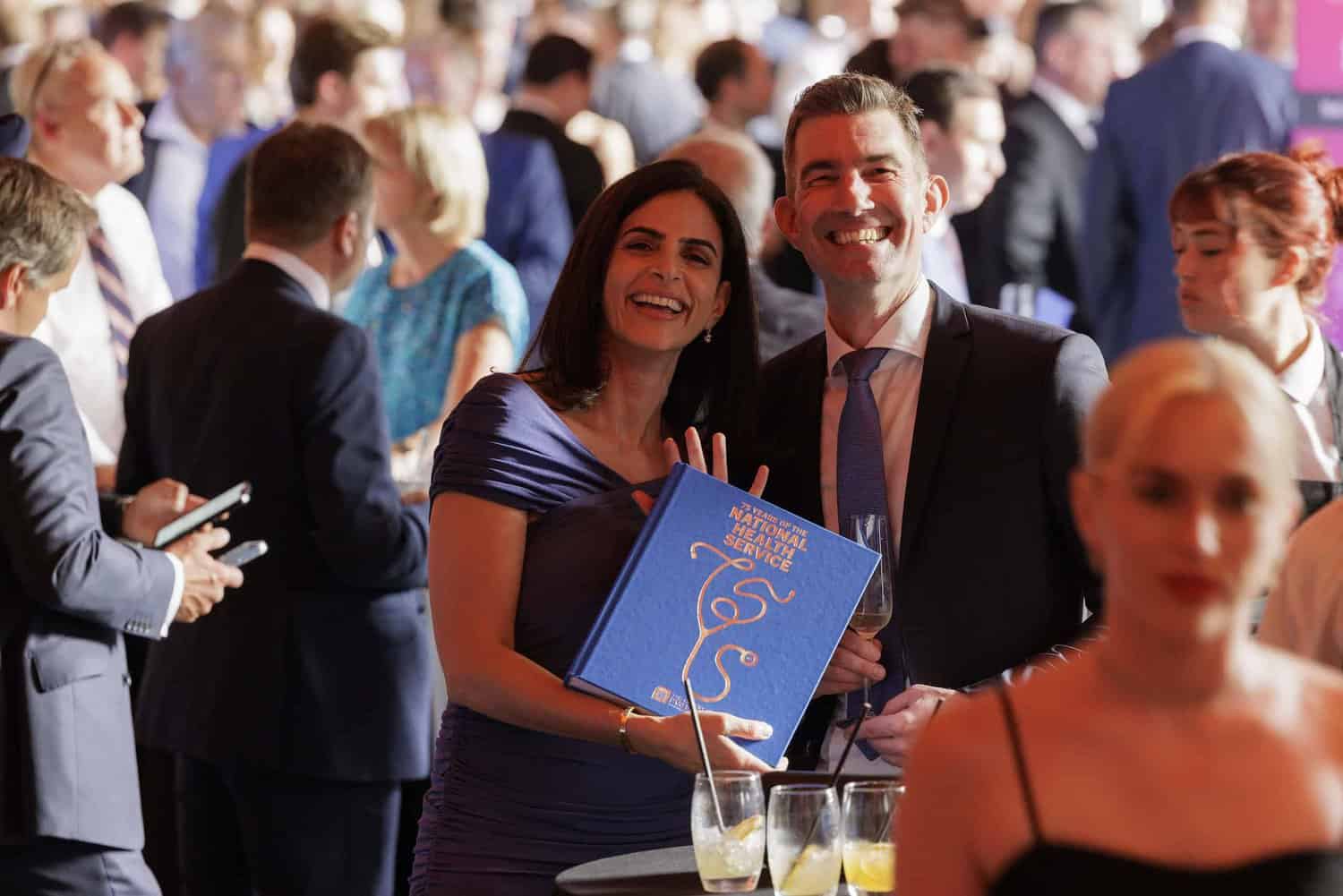 Two people smile at a crowded event, proudly holding a large blue book celebrating 75 years of the National Health Service. Others mingle in the background, sharing in the joy of this remarkable milestone.