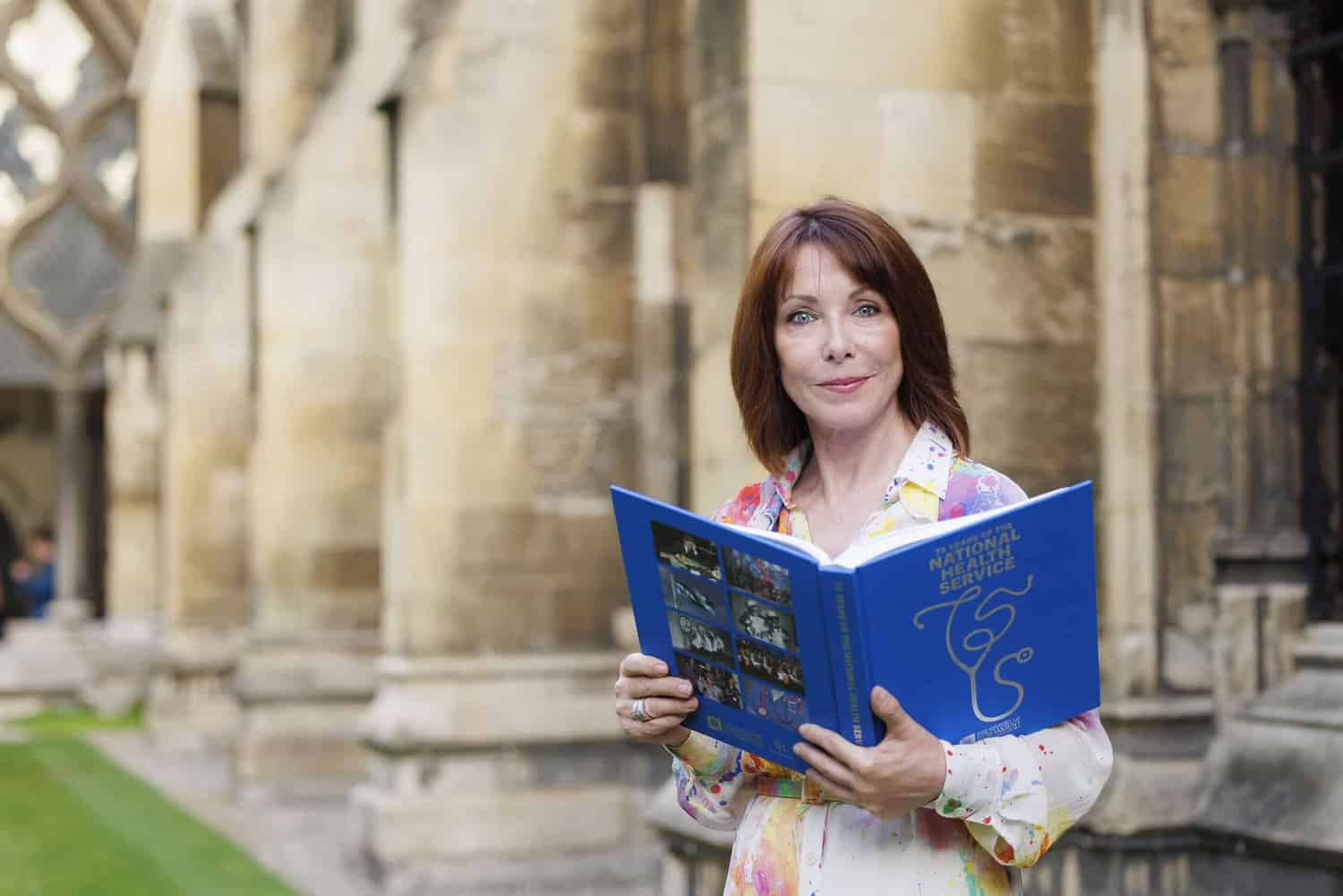 A person holding a blue book stands outside near a stone building, reflecting on 75 years of the National Health Service.