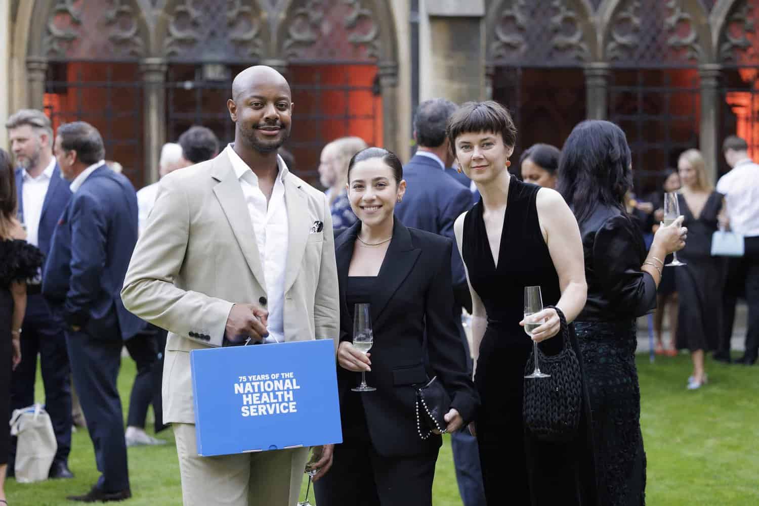 Three people holding drinks and a blue NHS sign stand outdoors at an event celebrating 75 years of the National Health Service. Others are mingling in the background, enjoying the festivities.