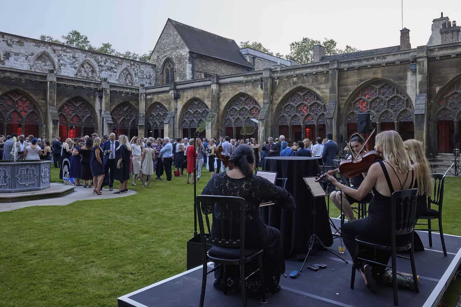 A string quartet performs at an outdoor event in a historic courtyard, celebrating 75 years of the National Health Service, surrounded by people dressed in formal attire.