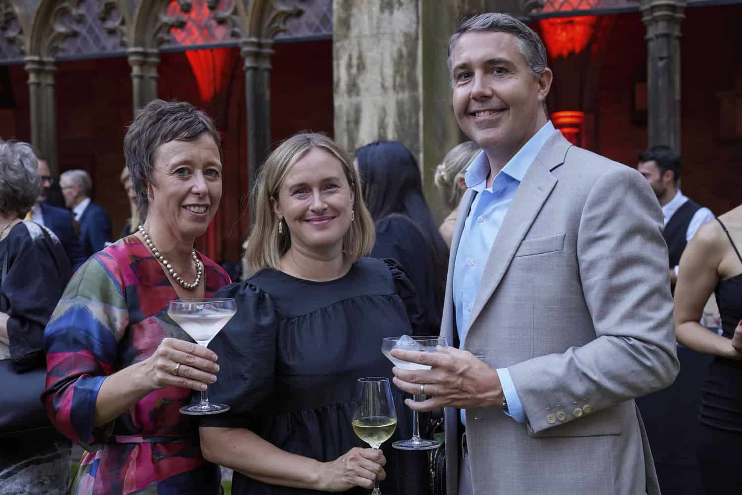 Three people holding drinks, standing together at an outdoor gathering celebrating the 75th NHS anniversary, dressed in formal attire. Medieval-style stone arches are visible in the background.