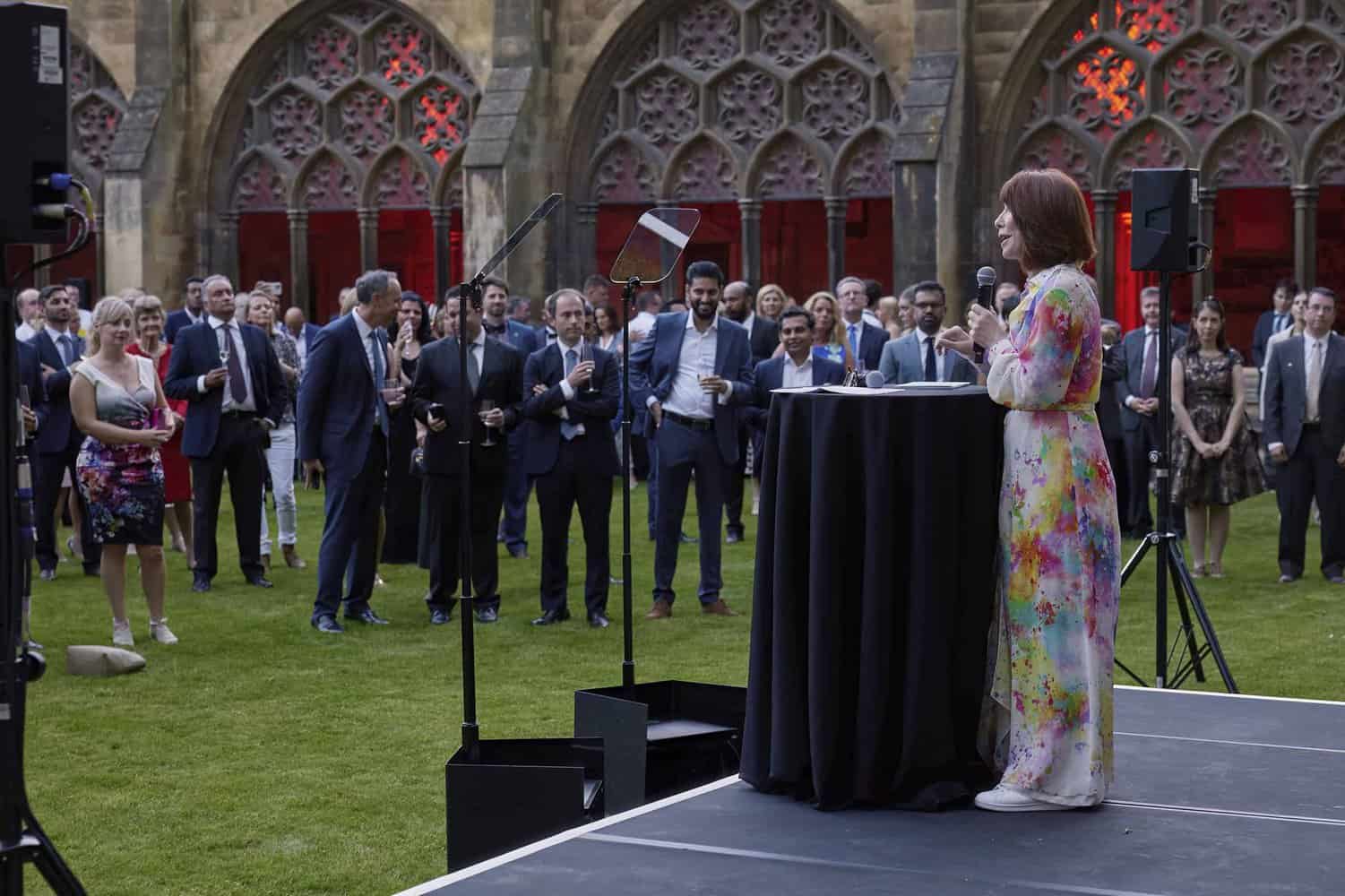 A woman in a colorful dress speaks at a podium to a well-dressed crowd gathered on a lawn, commemorating 75 years of the National Health Service, in front of a building with ornate arches.