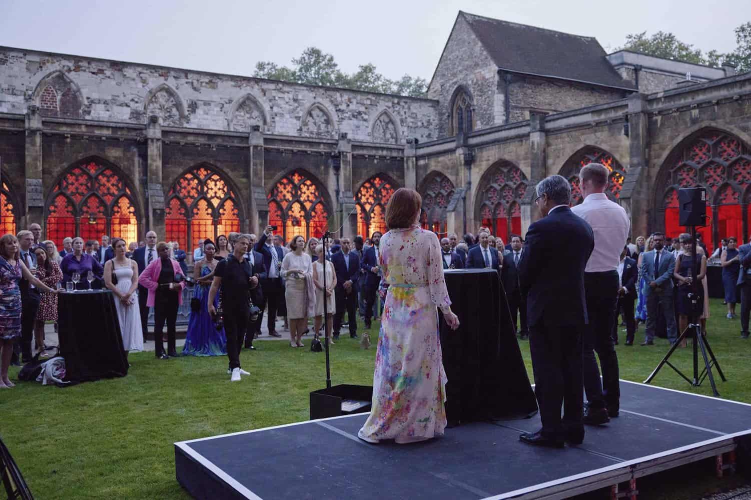 A group of people attends an outdoor event in a historic courtyard with red-illuminated arches, where three speakers stand on a raised platform, celebrating 75 years of the NHS.