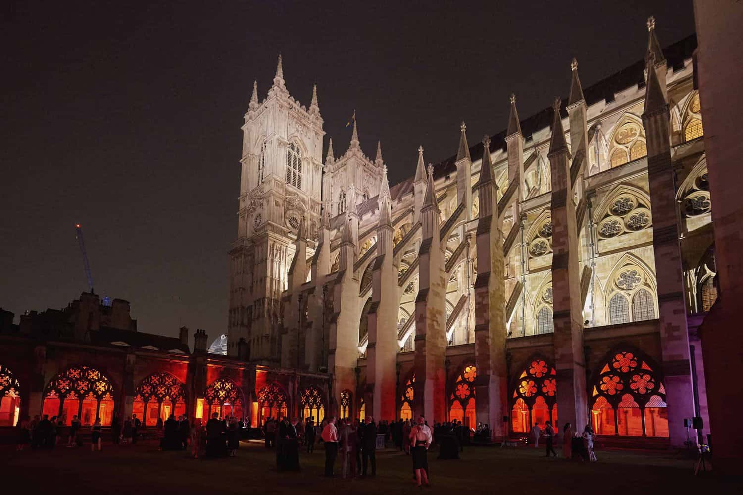 Exterior night view of an illuminated Gothic-style cathedral celebrates 75 years of the NHS, with people gathering in front. The scene is majestic, featuring pointed arches and intricate stained glass windows that honor the legacy and care the National Health Service has provided.