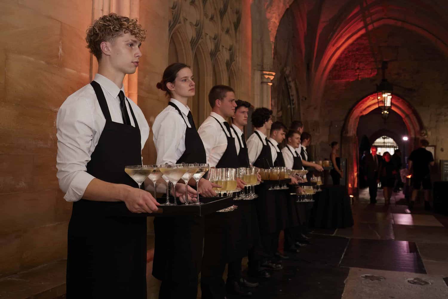 A row of servers in black aprons hold trays of cocktails in a dimly lit venue with arched architecture, celebrating 75 years since the founding of the National Health Service.