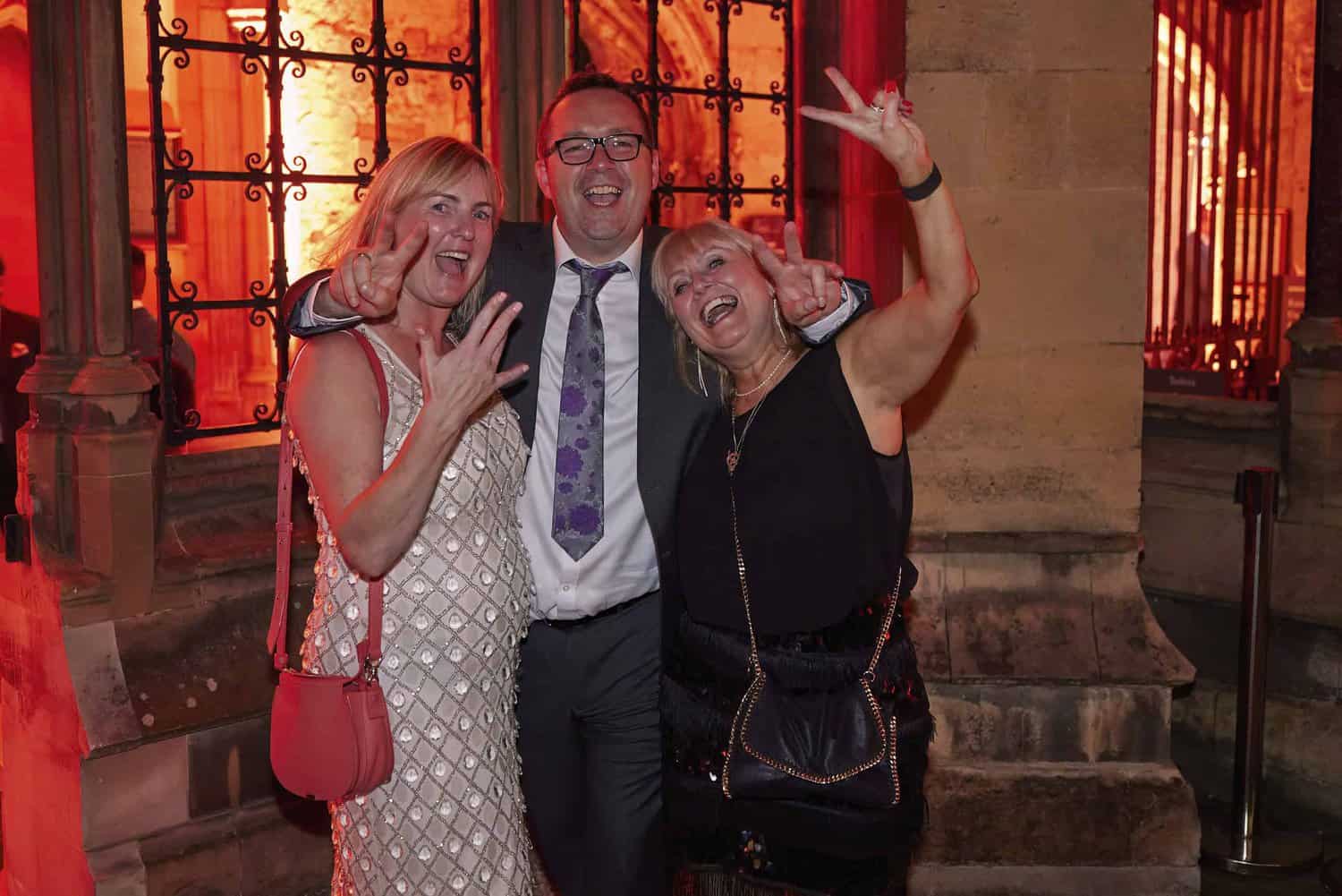 Three people, celebrating 75 years of the NHS, stand close together, smiling and making peace signs in front of a window with warm orange lighting.