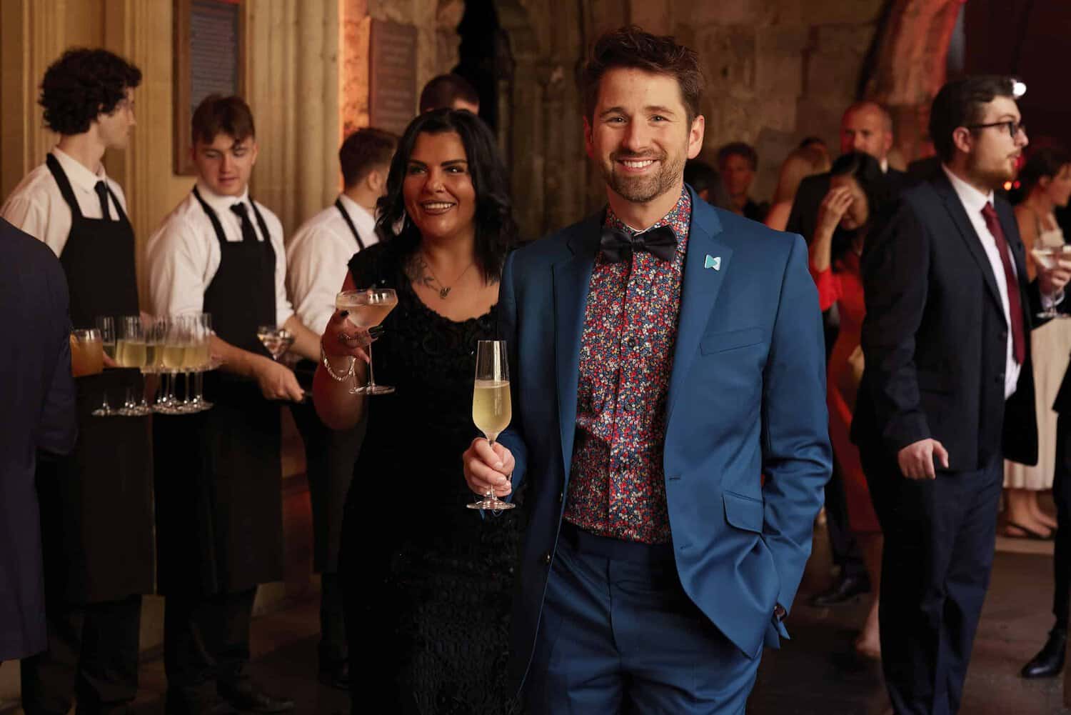 A man in a blue suit and a woman in a black dress celebrate 75 years of the National Health Service, holding drinks at the formal event, with waitstaff and guests visible in the background.