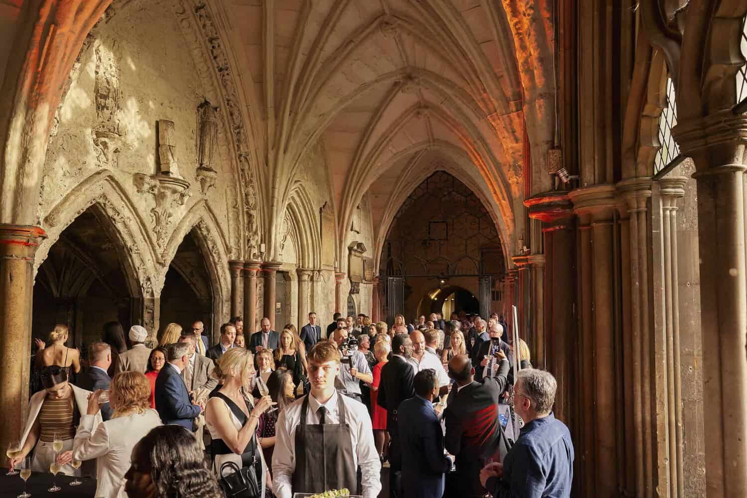 A large group of people is socializing in a historic, arched hallway with stone columns and vaulted ceilings, celebrating 75 years of the National Health Service.