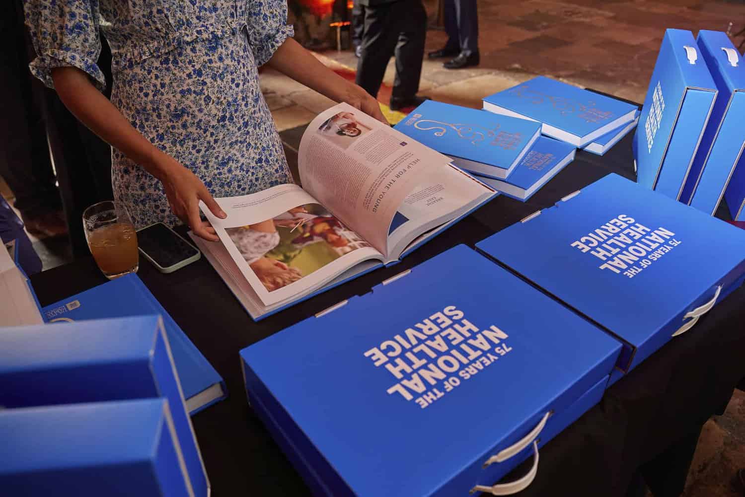 A person in a floral top gazes at an open book on a table surrounded by vibrant blue bags celebrating "75 Years of the NHS.