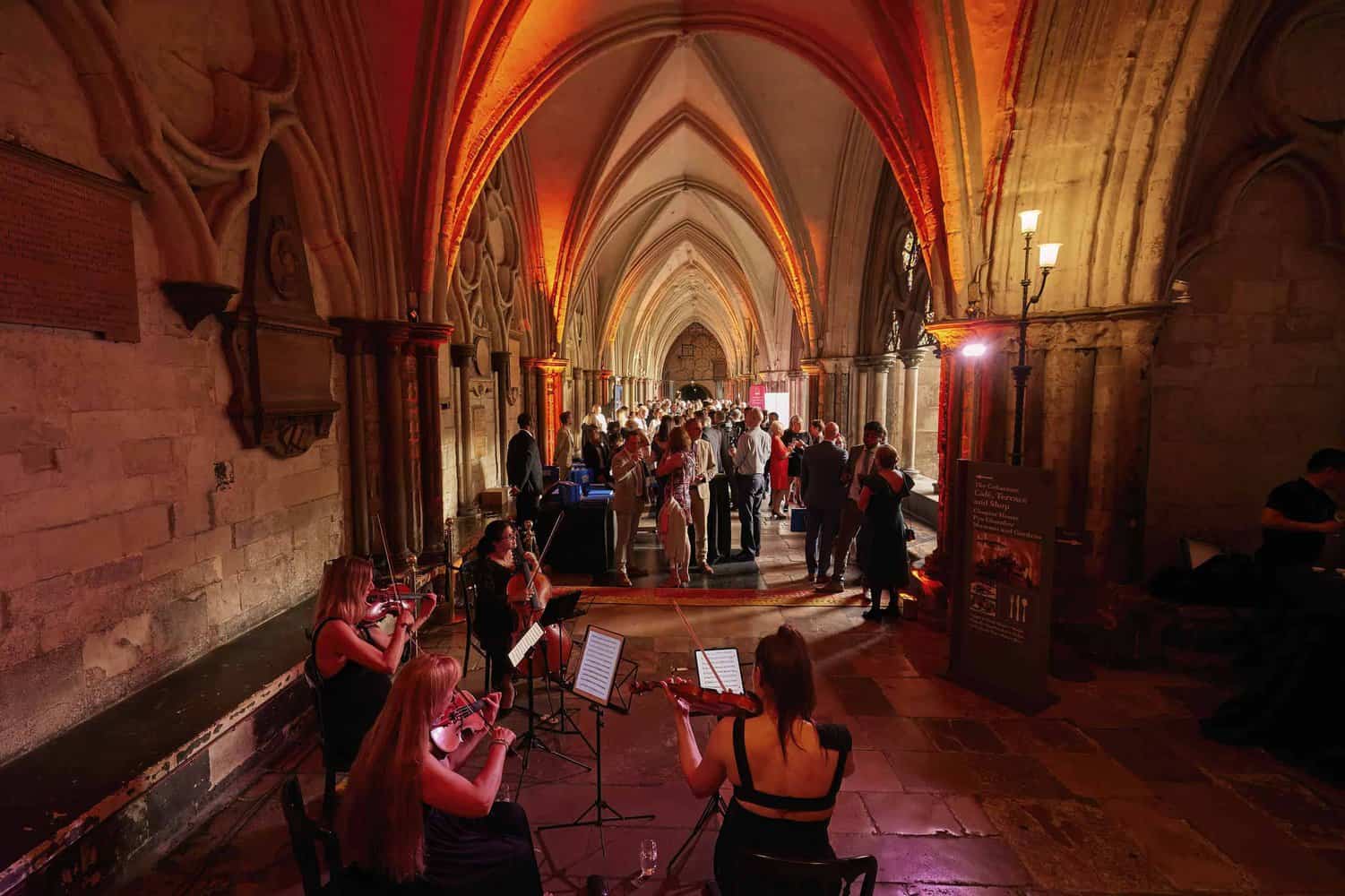Musicians play string instruments in a historic church hallway with arched ceilings, celebrating 75 years of the NHS as a crowd gathers in the background under warm lighting.