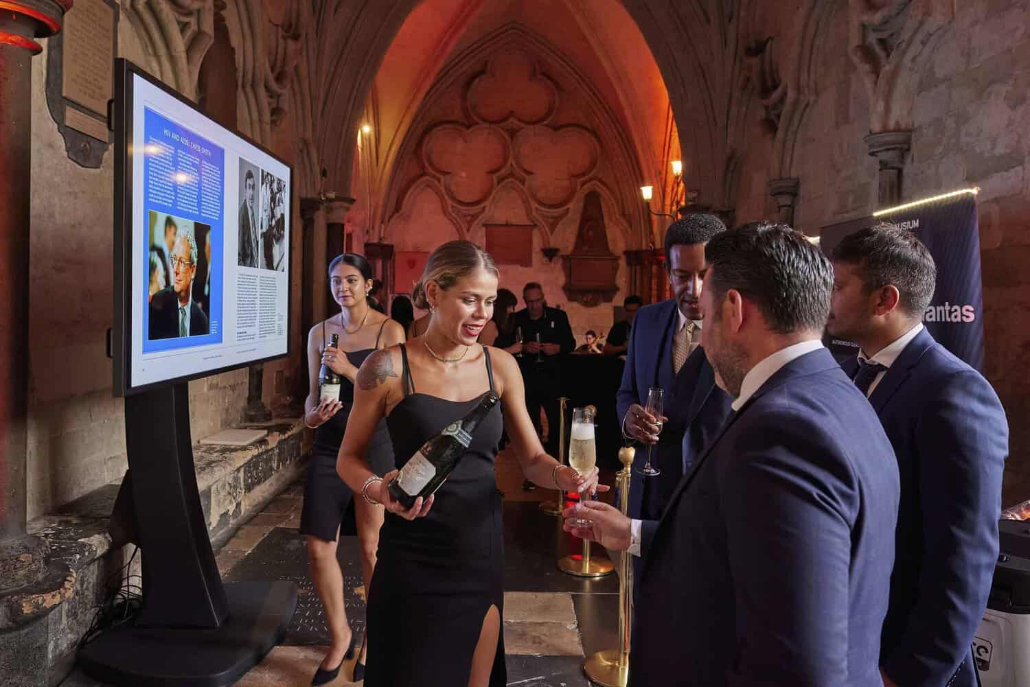 A woman in a black dress showcases a wine bottle to four people in formal attire at an indoor celebration. A large screen highlights images and text celebrating 75 years of the National Health Service in the background.