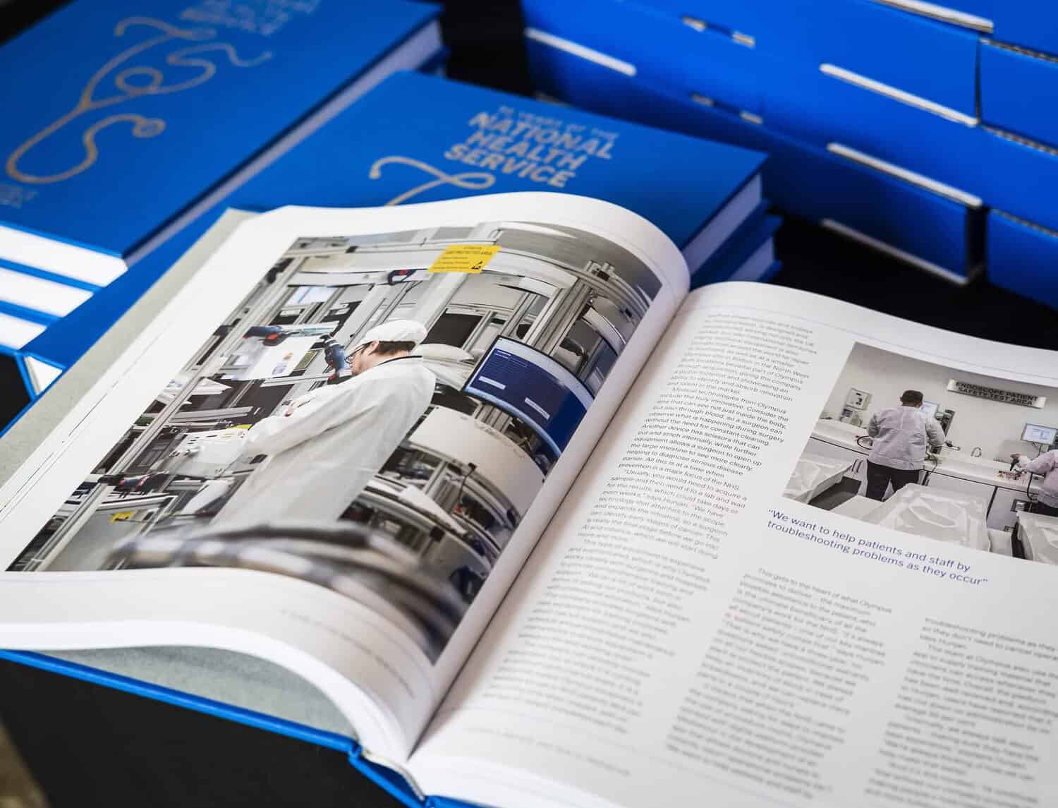 Open book on a table next to stacked blue booklets. The book displays an image of a person in a laboratory and a page of text, commemorating 75 years of the National Health Service's remarkable journey.