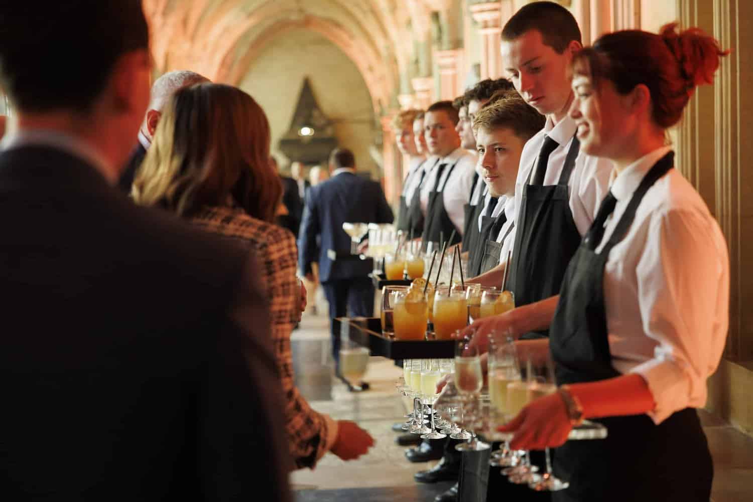 Servers in black aprons line the hallway, offering drinks to guests celebrating 75 years of the NHS, their trays brimming with refreshing beverages at this special event.