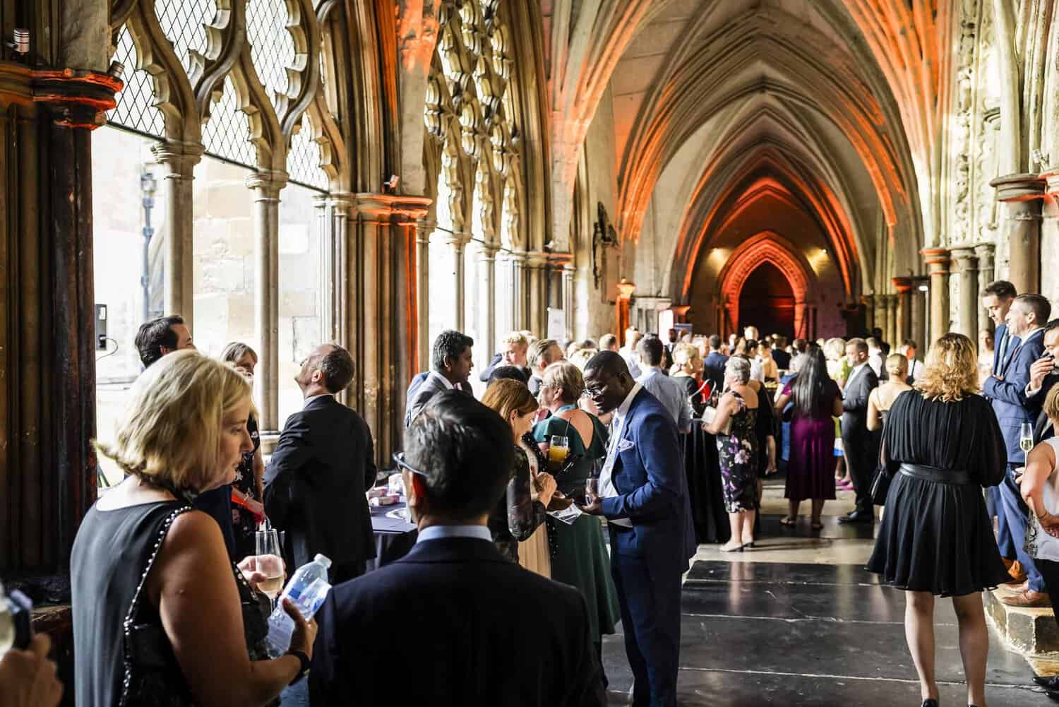 A group of people in formal attire gathered in a cathedral-like hallway with arched ceilings and ornate columns, celebrating 75 years of the National Health Service while engaged in conversation and socializing.