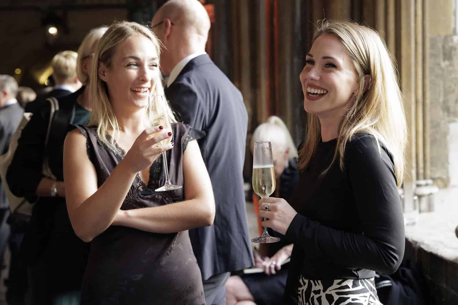 Two women in conversation hold champagne glasses at a social gathering celebrating 75 years of the NHS. People in formal attire are in the background, adding to the celebratory atmosphere.