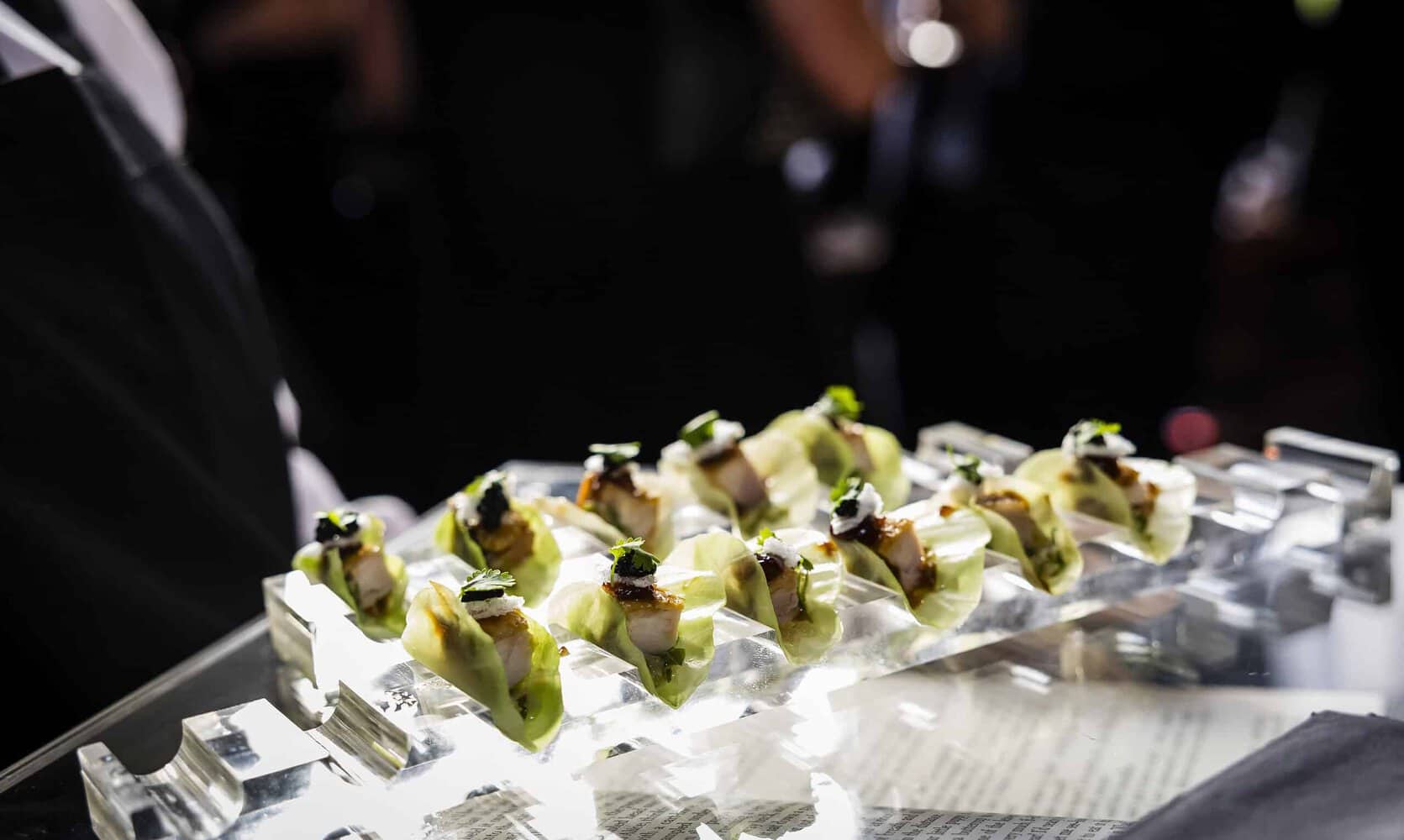 A tray with neatly arranged hors d'oeuvres featuring avocado and other garnishes on a reflective surface celebrates 75 years of the NHS, set against a blurred, dark background.