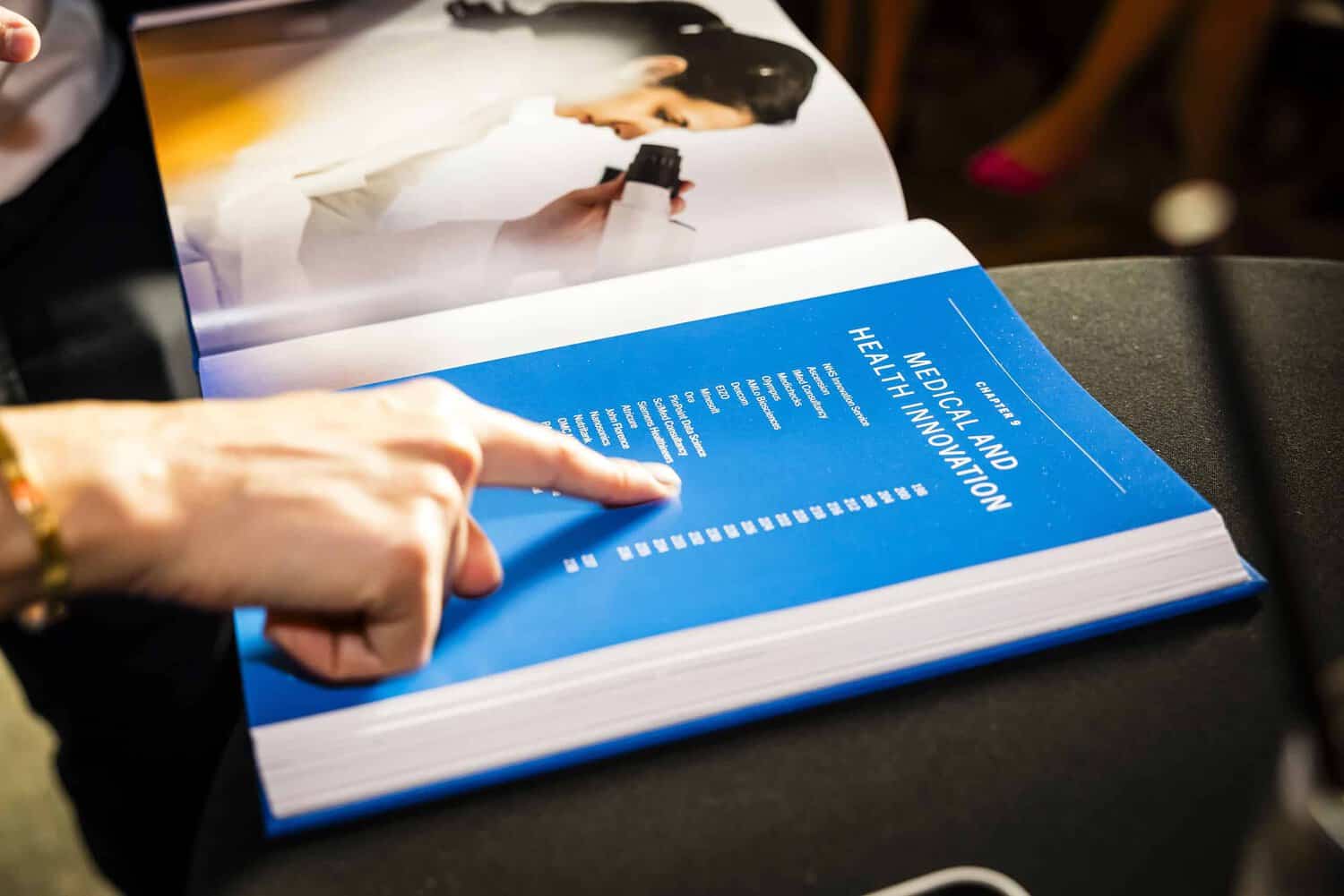 A person points at a blue book page titled "Medical and Health Innovation," with a blurred open page featuring a woman in the background, celebrating 75 years of the National Health Service's groundbreaking contributions to healthcare.