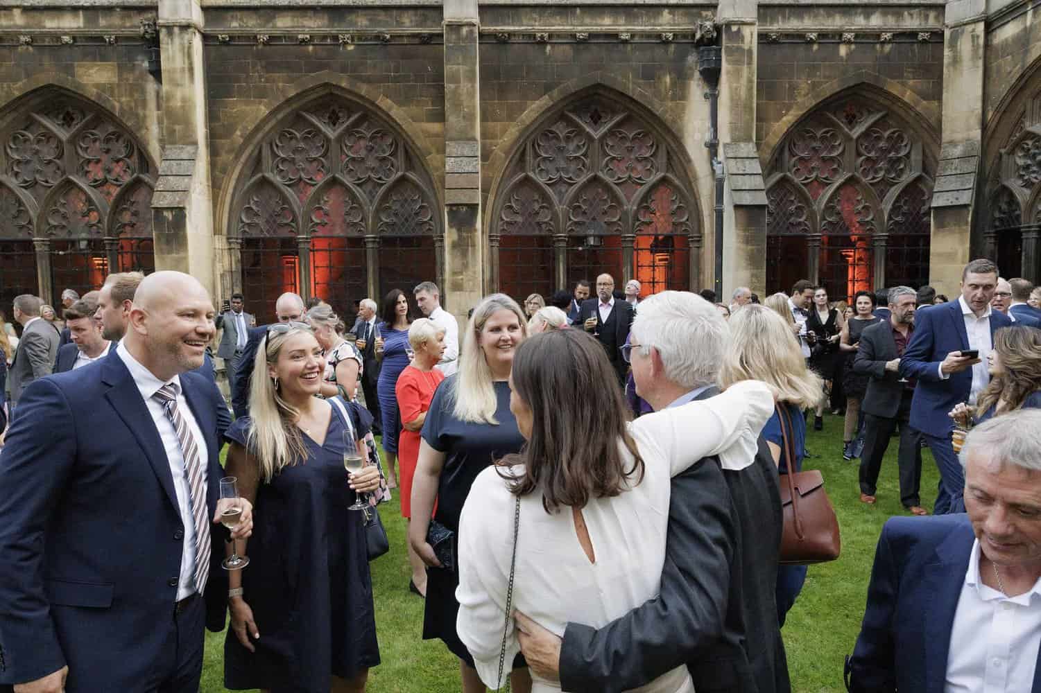 A group of people in formal attire mingles in a courtyard with Gothic architecture, celebrating 75 years of the National Health Service.