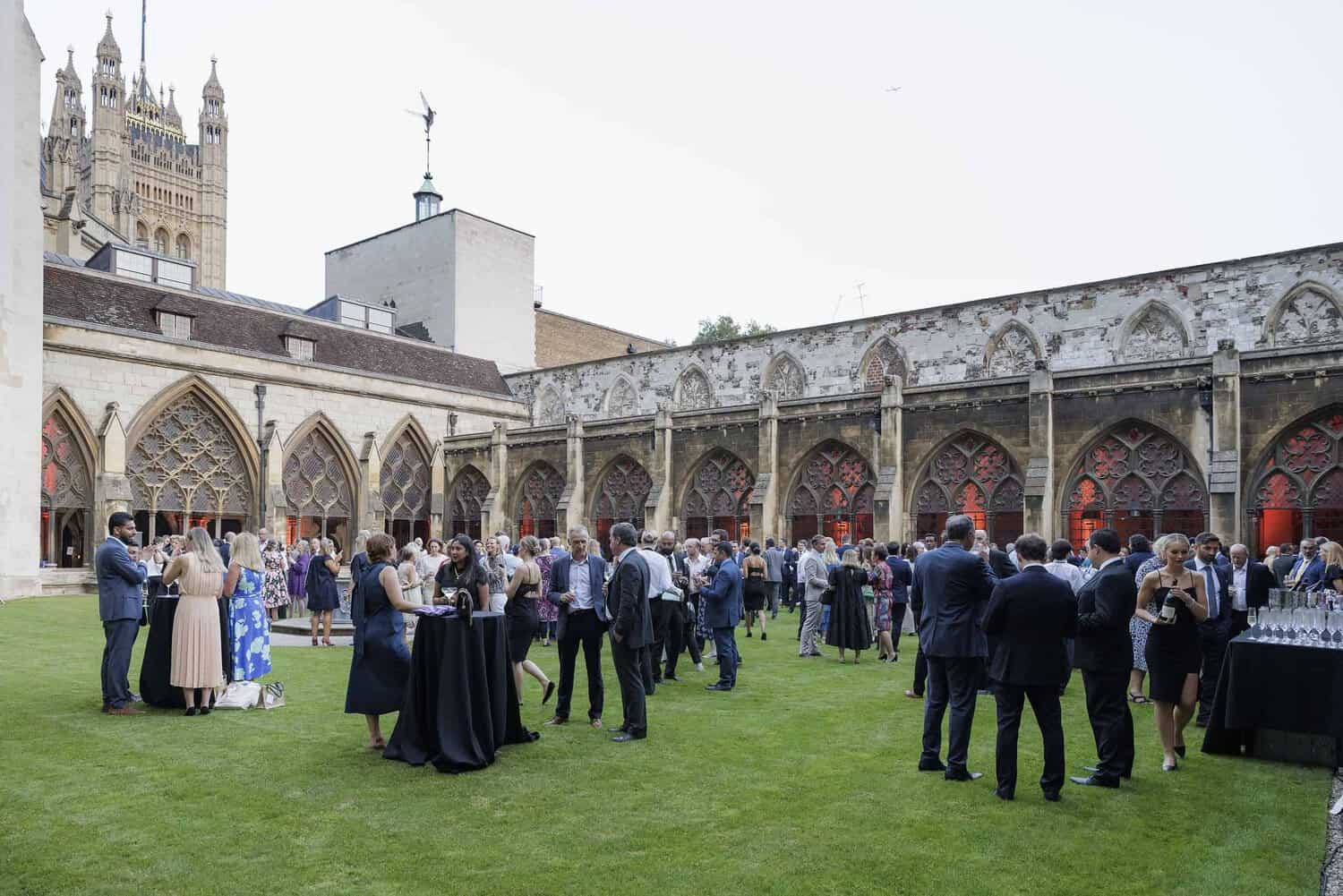 A large group of people is mingling at an outdoor event in a courtyard adorned with Gothic architecture, celebrating 75 years of the National Health Service.