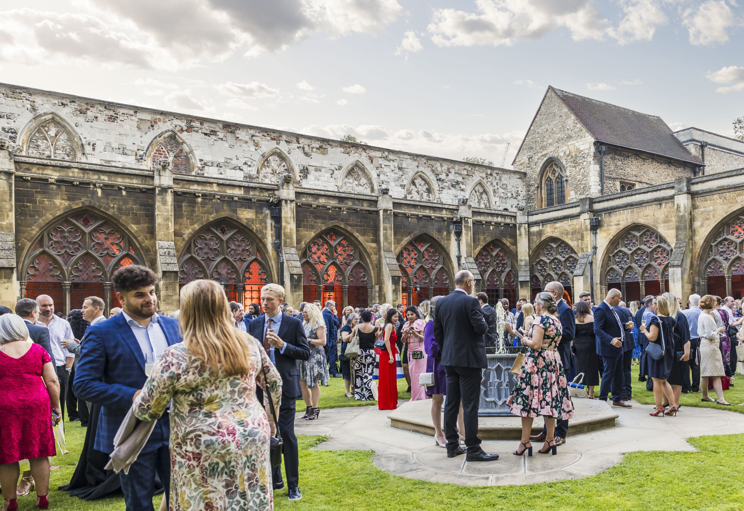 People in formal attire are socializing at an outdoor event in a historic building courtyard, celebrating 75 years of the NHS amidst arched windows and stone walls.