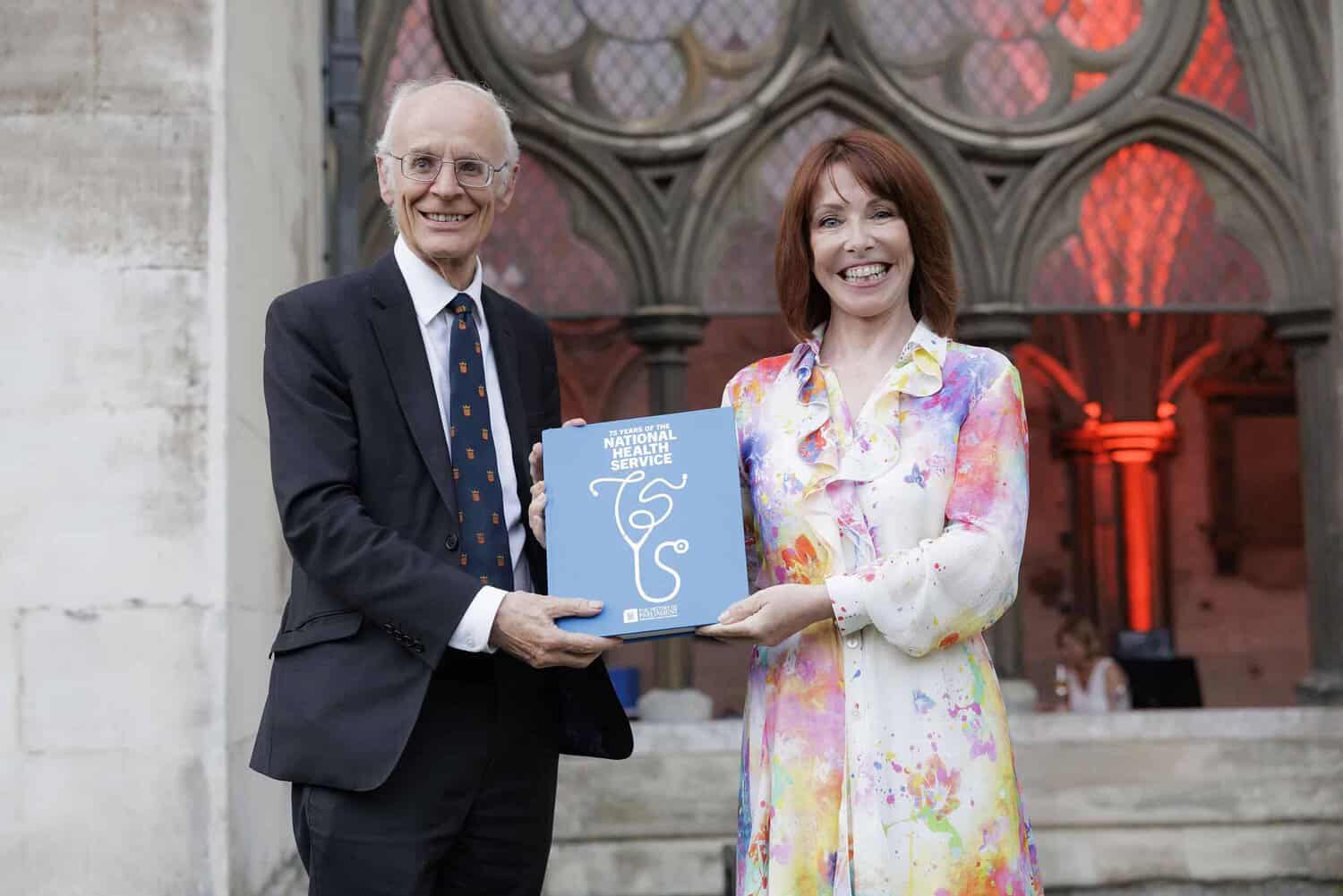 Two people stand side by side outside a building, each holding a side of a book titled "75 Years of the NHS". They are smiling and dressed formally.