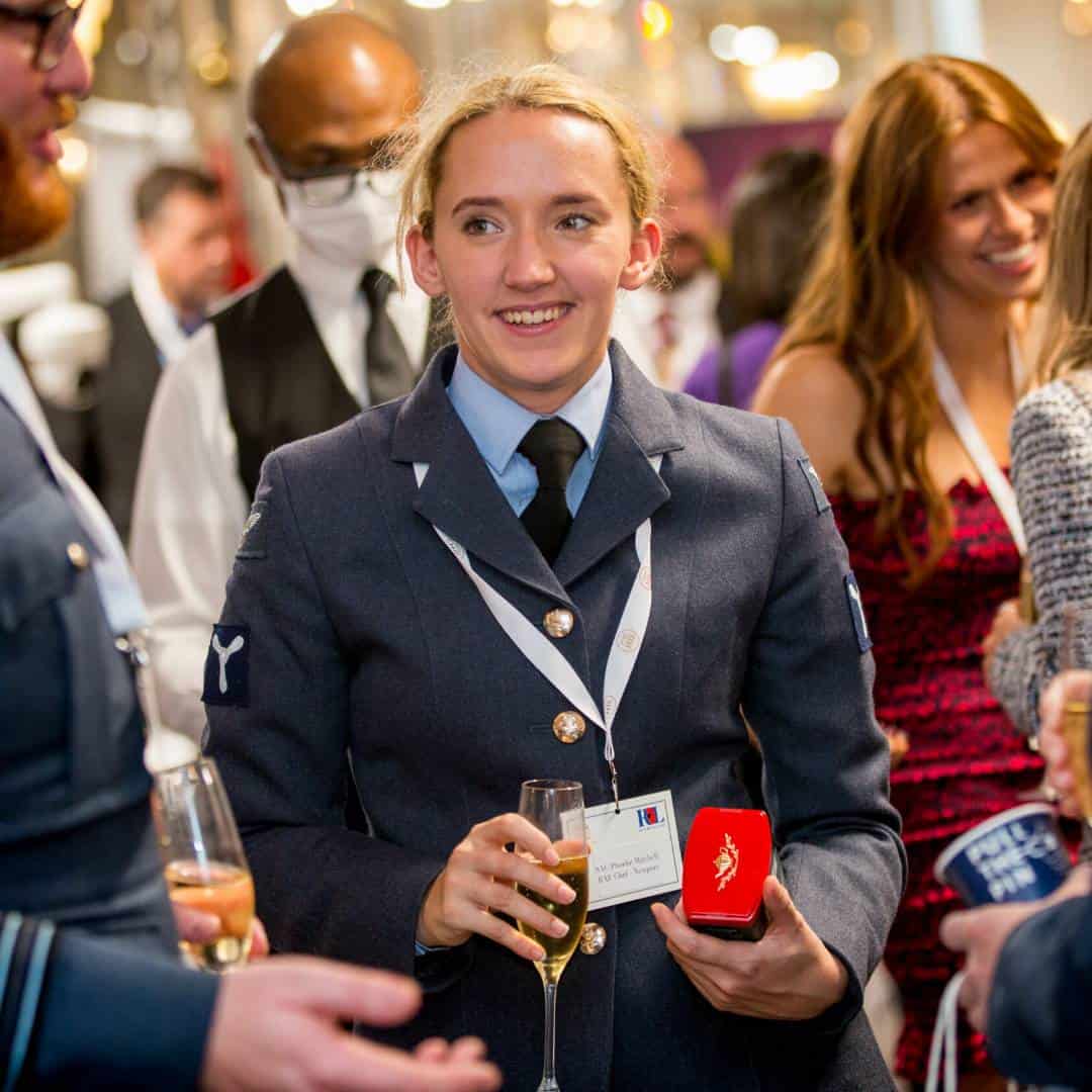 A person in a military uniform holds a drink and an award at a social event celebrating heroes, surrounded by people, including someone in a red dress and a man with a mask.