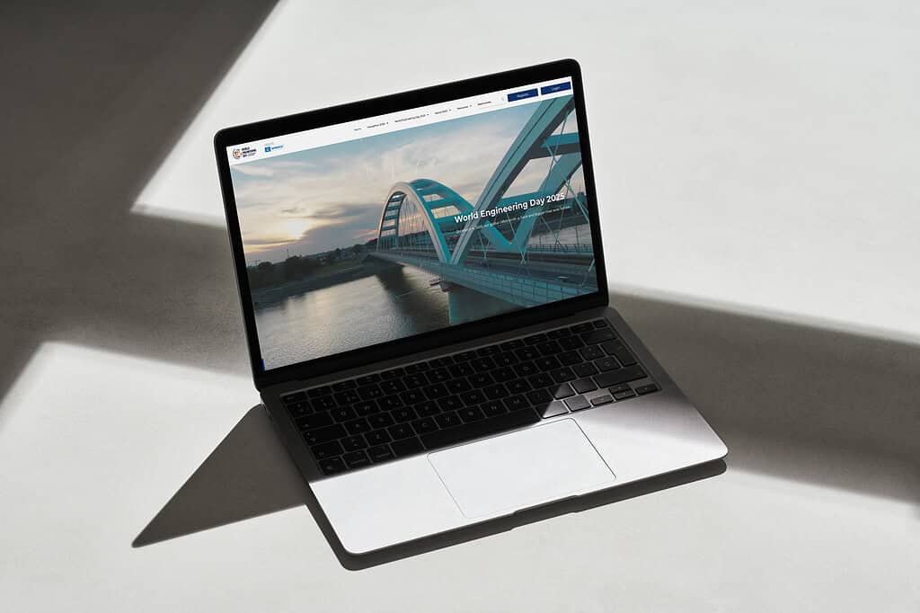 A laptop on a surface displays a website with a bridge image, emphasizing "World Engineering Day 2021" as part of the global celebration. Sunlight creates geometric shadows across the surface and device, adding an artistic touch to this ode to engineering achievements.