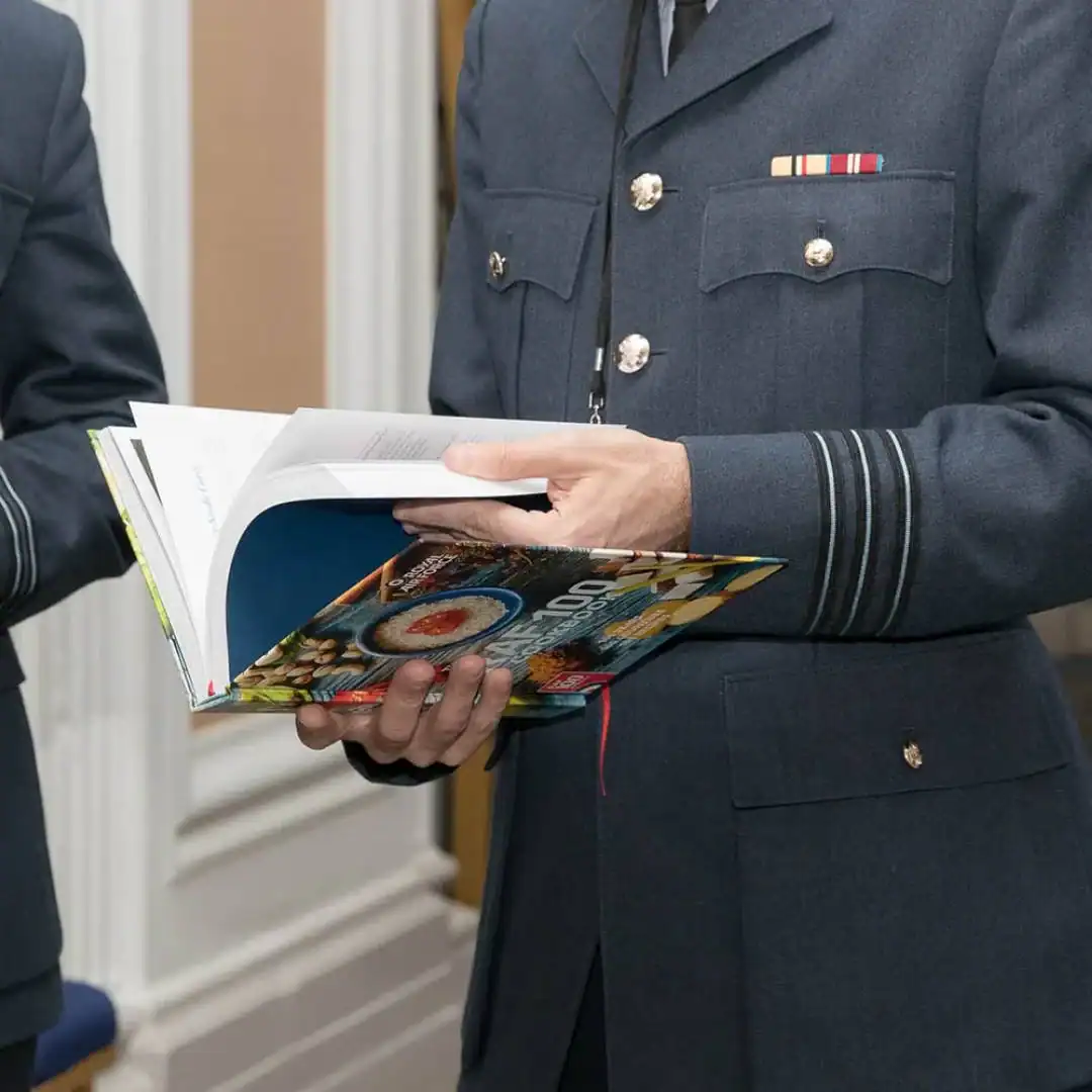 A person in a formal military uniform is indoors, holding and reading an open cookbook, perhaps discovering new culinary creations as part of the RAF 100 celebrations.
